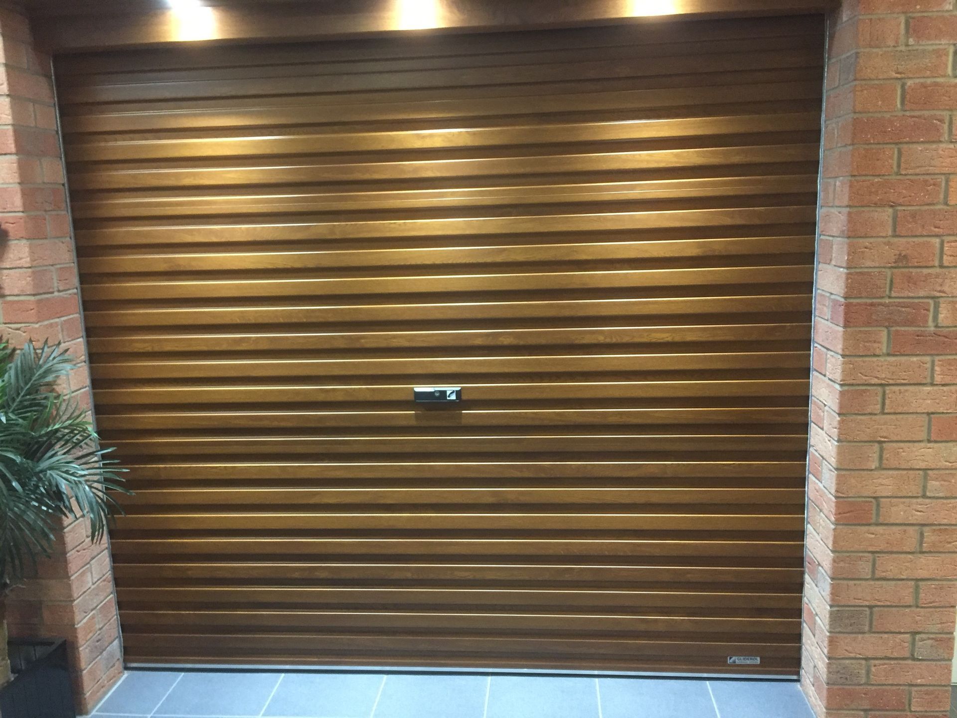 A brown, slatted roller garage door installed between two brick pillars, featuring a central lock and overhead lighting.