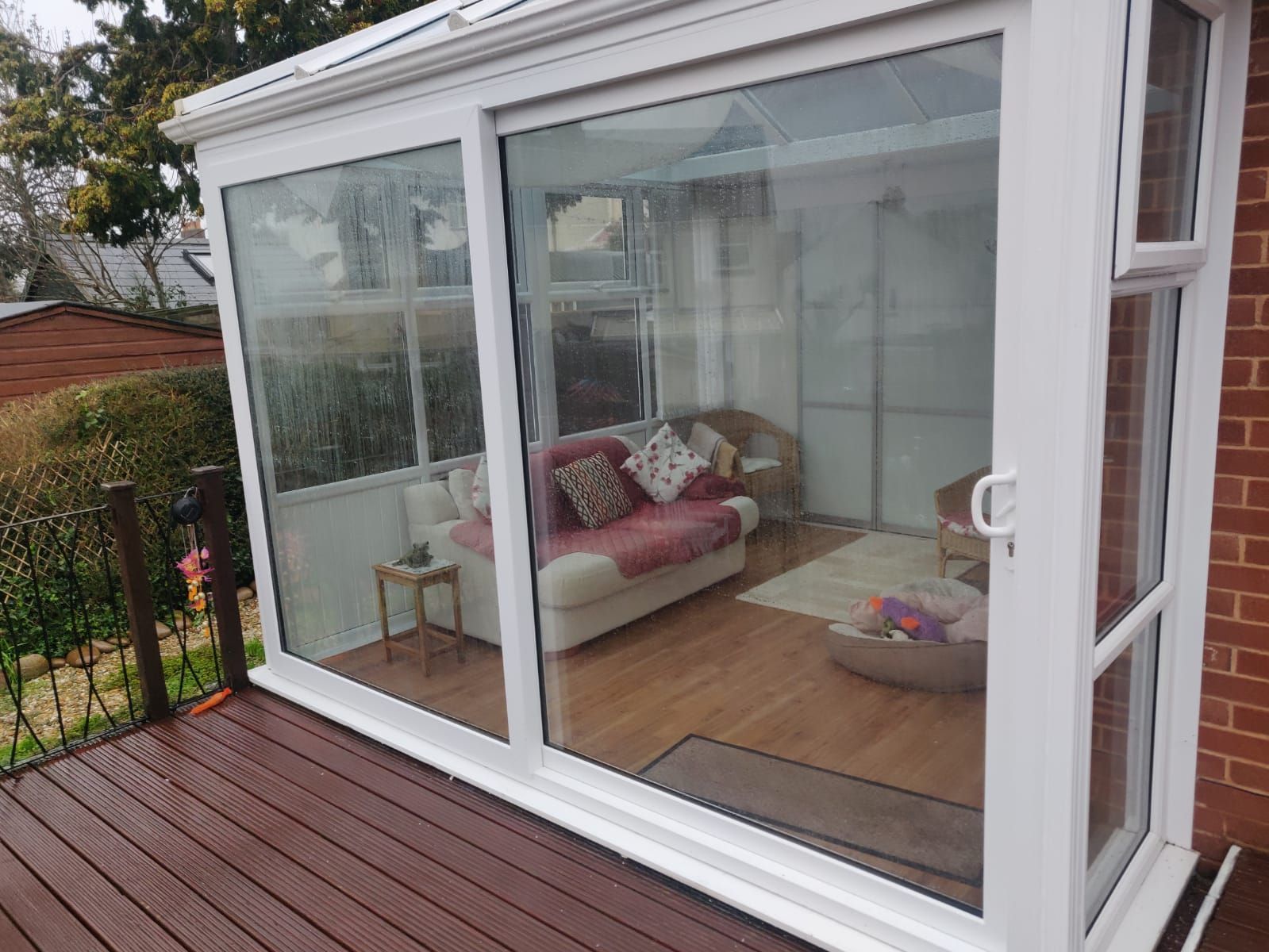 A white-framed conservatory with sliding doors, overlooking a wooden deck with a sofa and furniture visible inside.