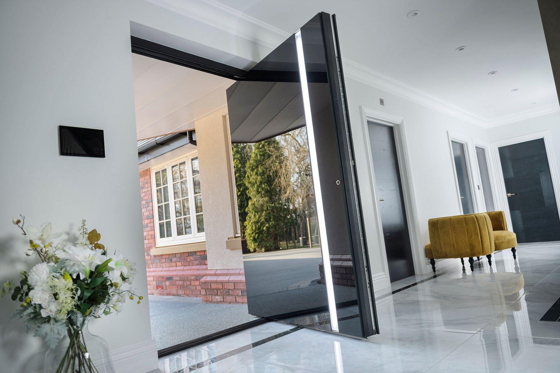 A modern home entrance with a large, black pivoting door slightly open, revealing an exterior view, in a bright hallway.