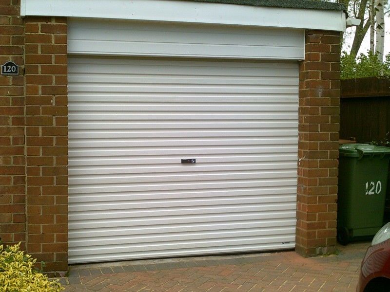 A closed white roller garage door on a brick building with a green wheelie bin next to it.