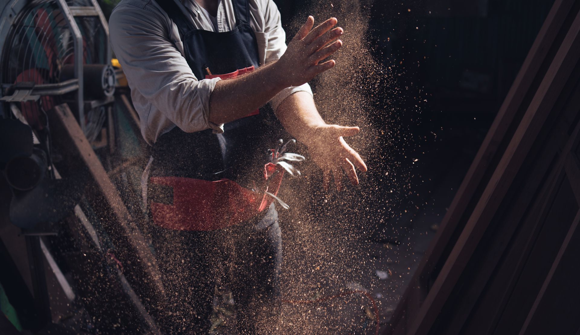Person clapping hands, releasing sawdust into the air. Workshop setting, with wood pieces.