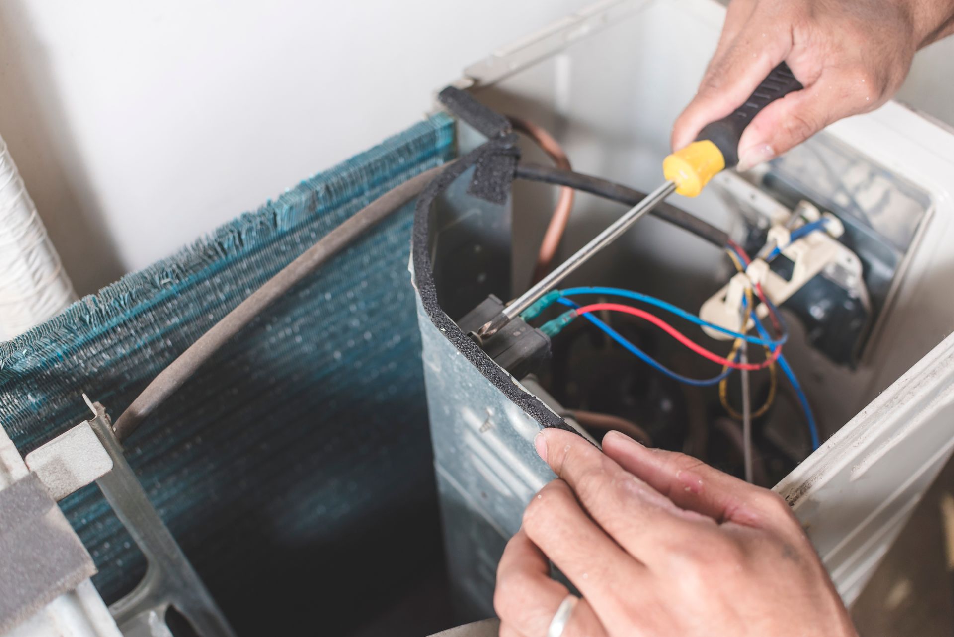 Hands using screwdriver to repair AC unit, working with wires and components.