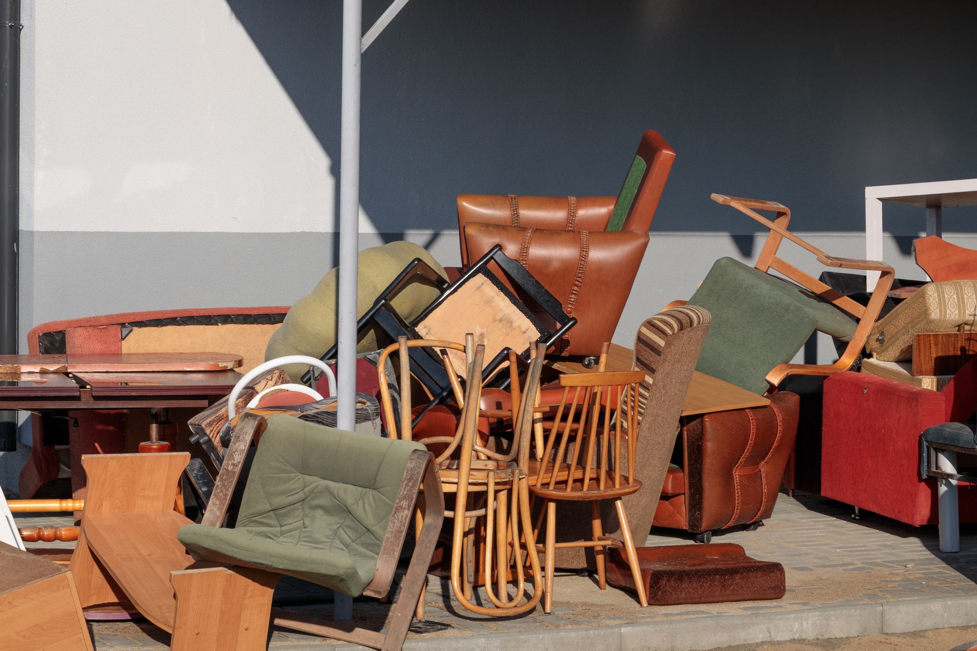 Pile of discarded furniture: chairs, tables, and upholstery, outside a building in sunlight.
