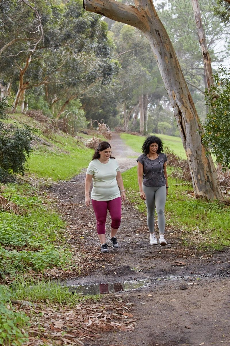 Two women walking in exercise gear along a bush path with grass and trees around