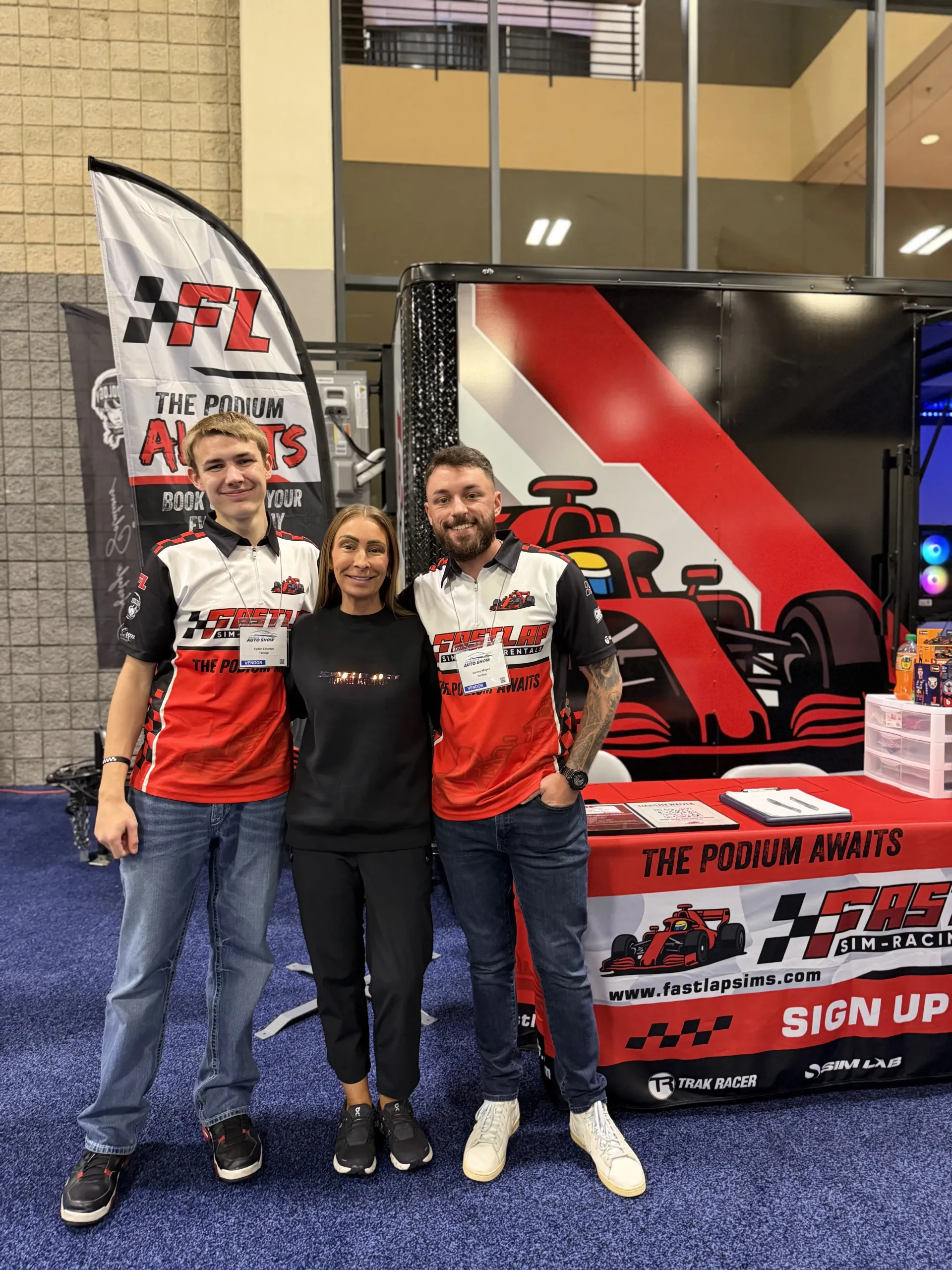 Three people posing at a motorsports booth with a race car display and a red-and-white sign-up banner