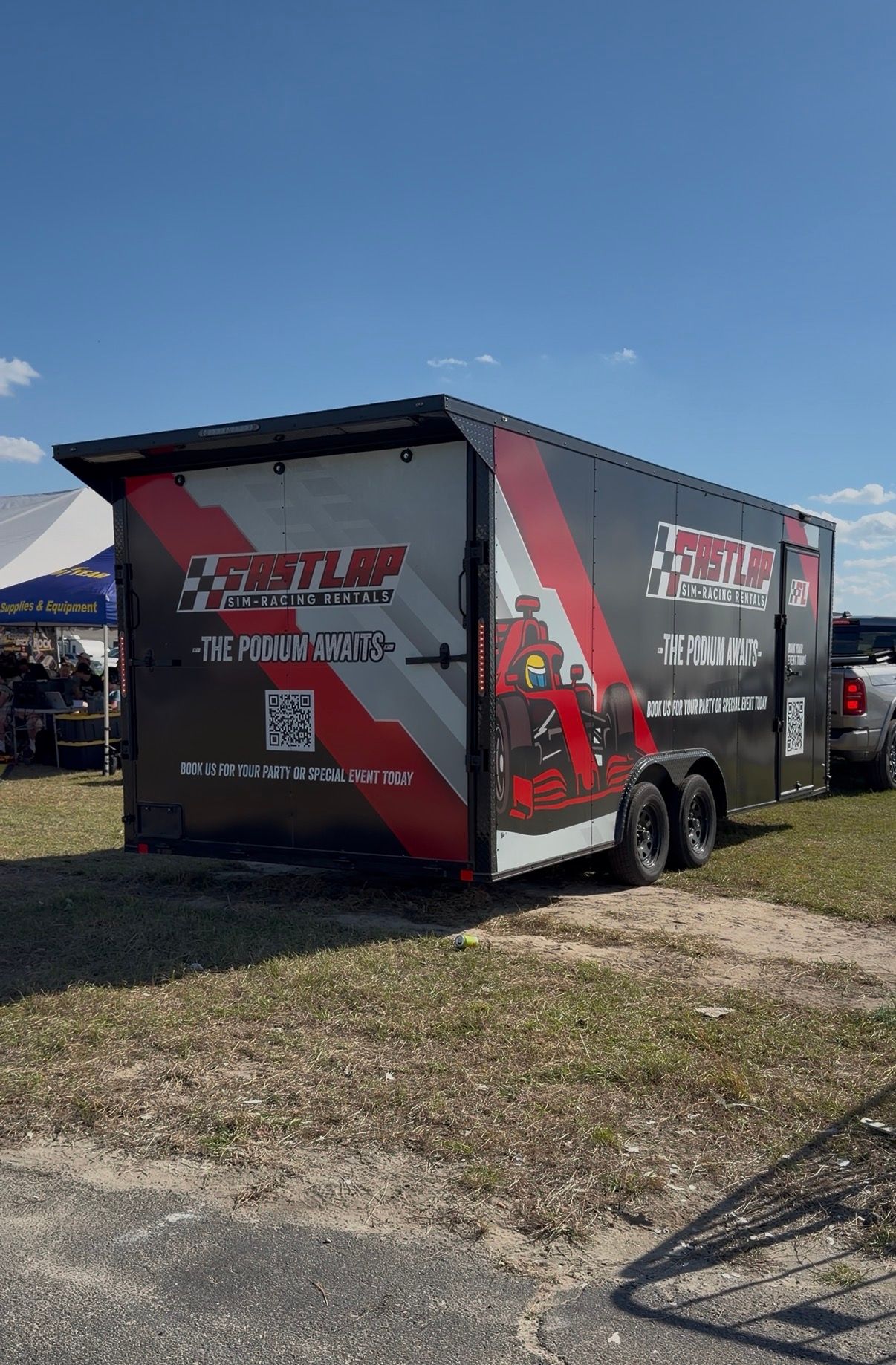 Red and black enclosed trailer with racing graphics parked on a grassy lot under a blue sky.