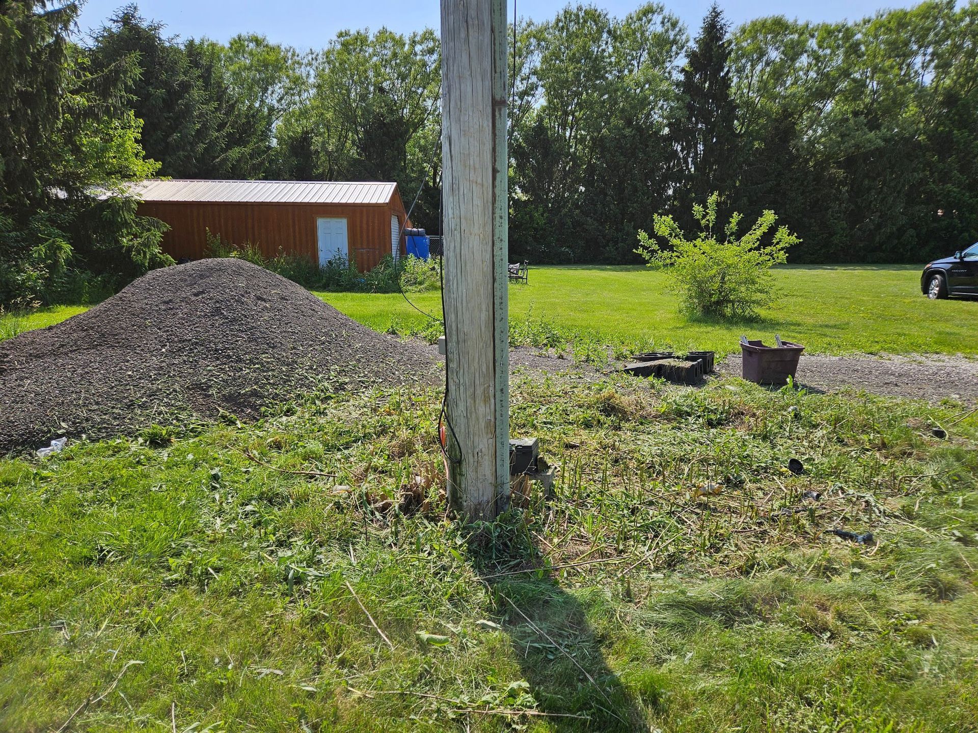 Gravel pile, utility pole, grassy yard, shed in the background under blue sky.