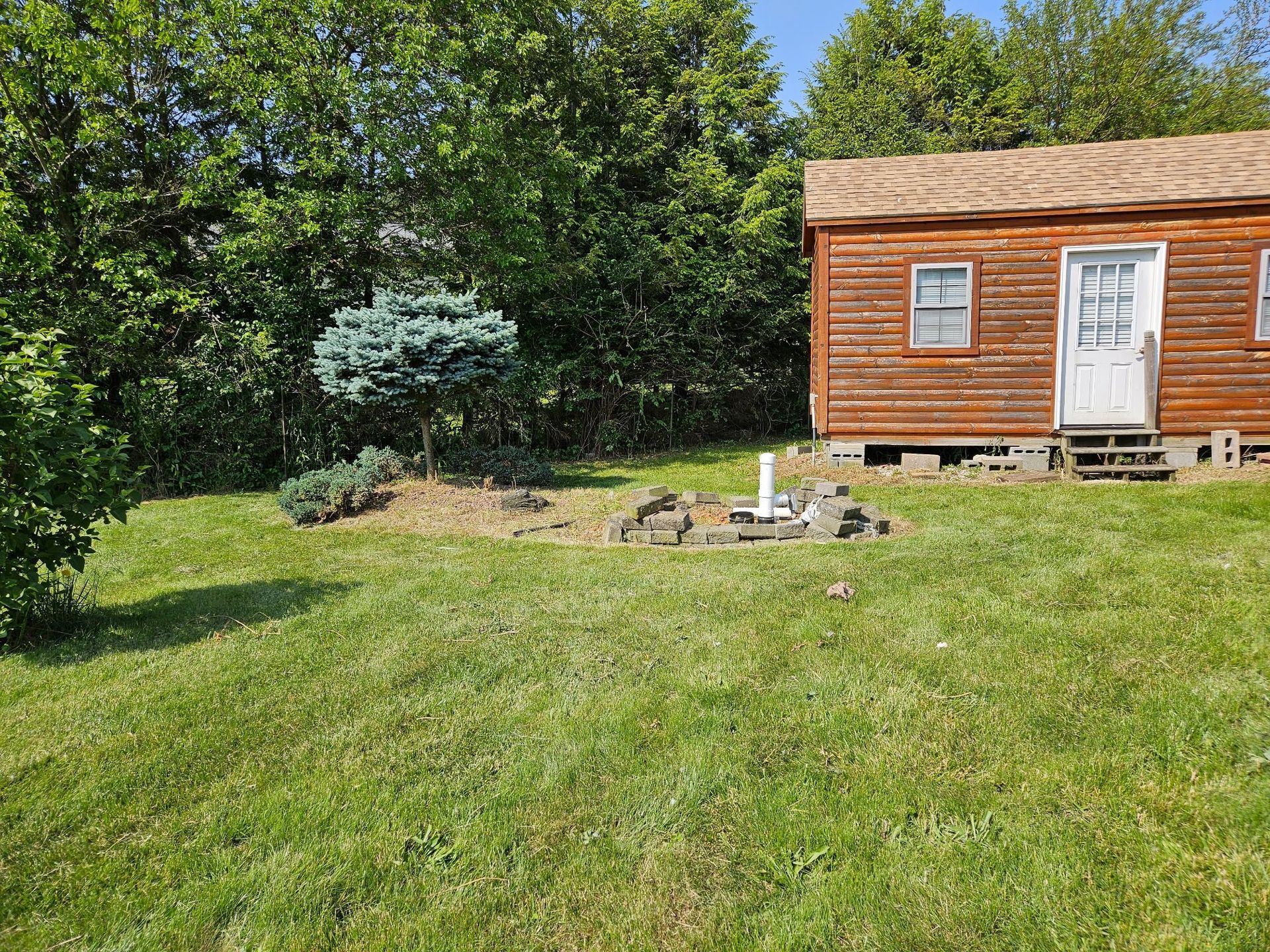 Wooden cabin with a blue spruce tree and a small garden, set on a grassy lawn.