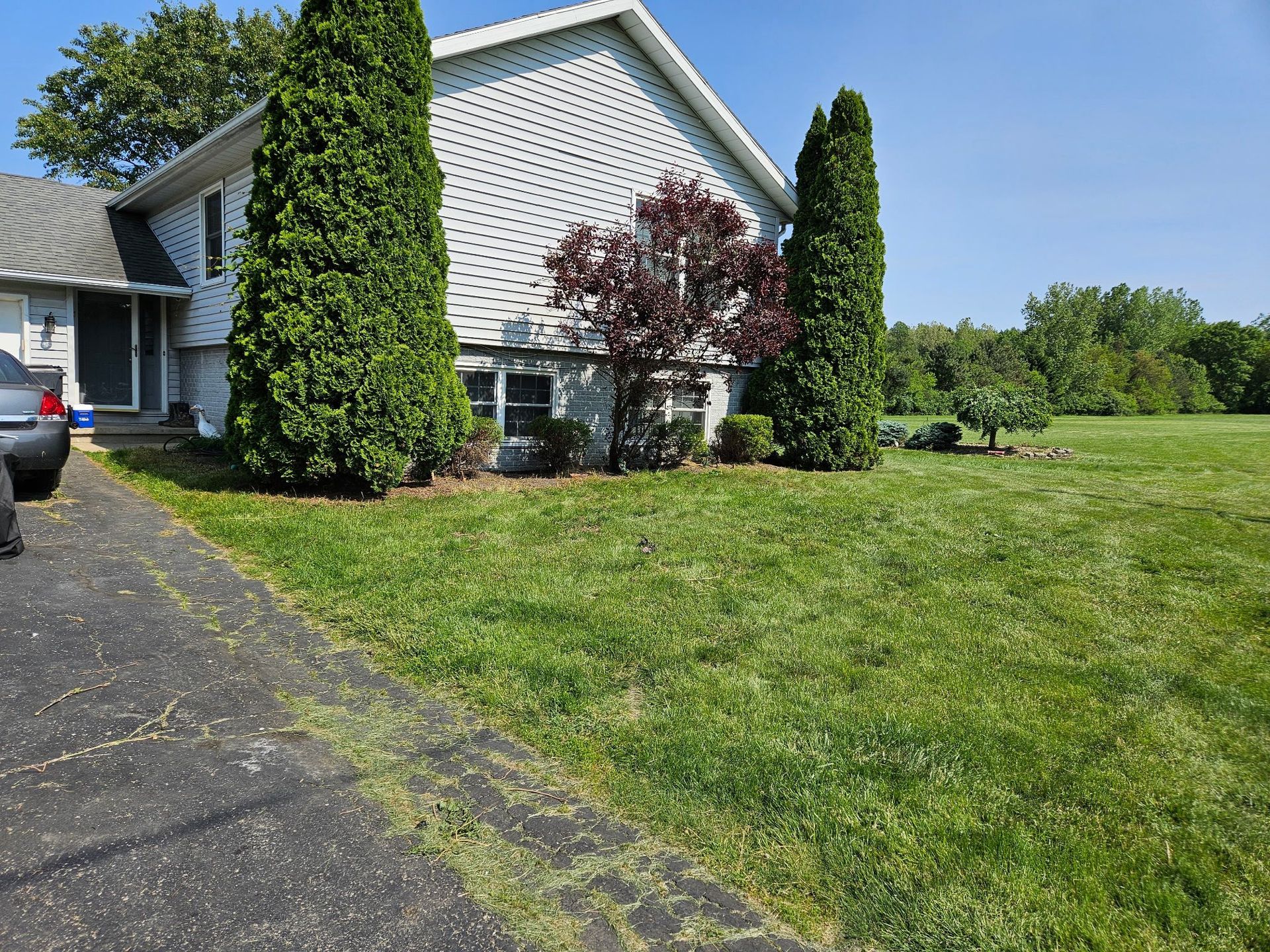 House with a green lawn, driveway, and trees. Blue sky in the background.
