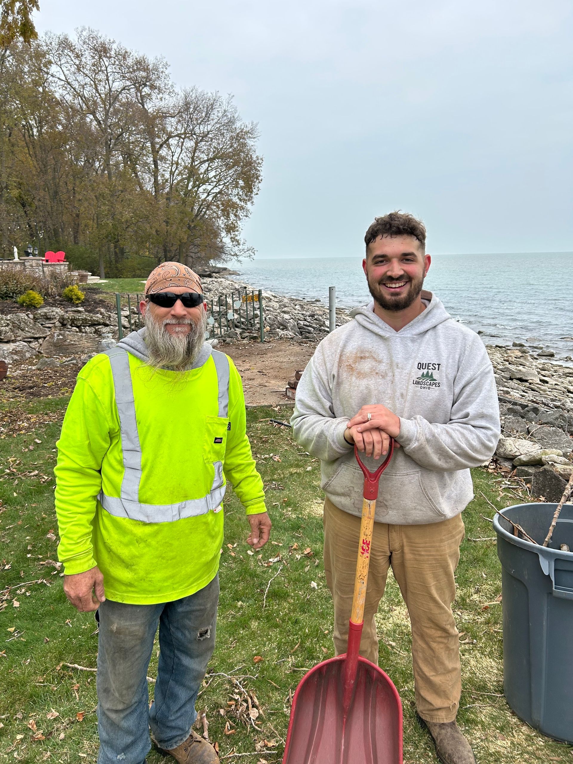 Two men stand on grassy shore, one holds shovel. Overcast sky, lake in background.