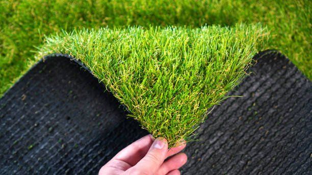 A hand holds up a corner of a square of artificial grass in Raleigh, revealing the dark, textured backing against a lawn background.
