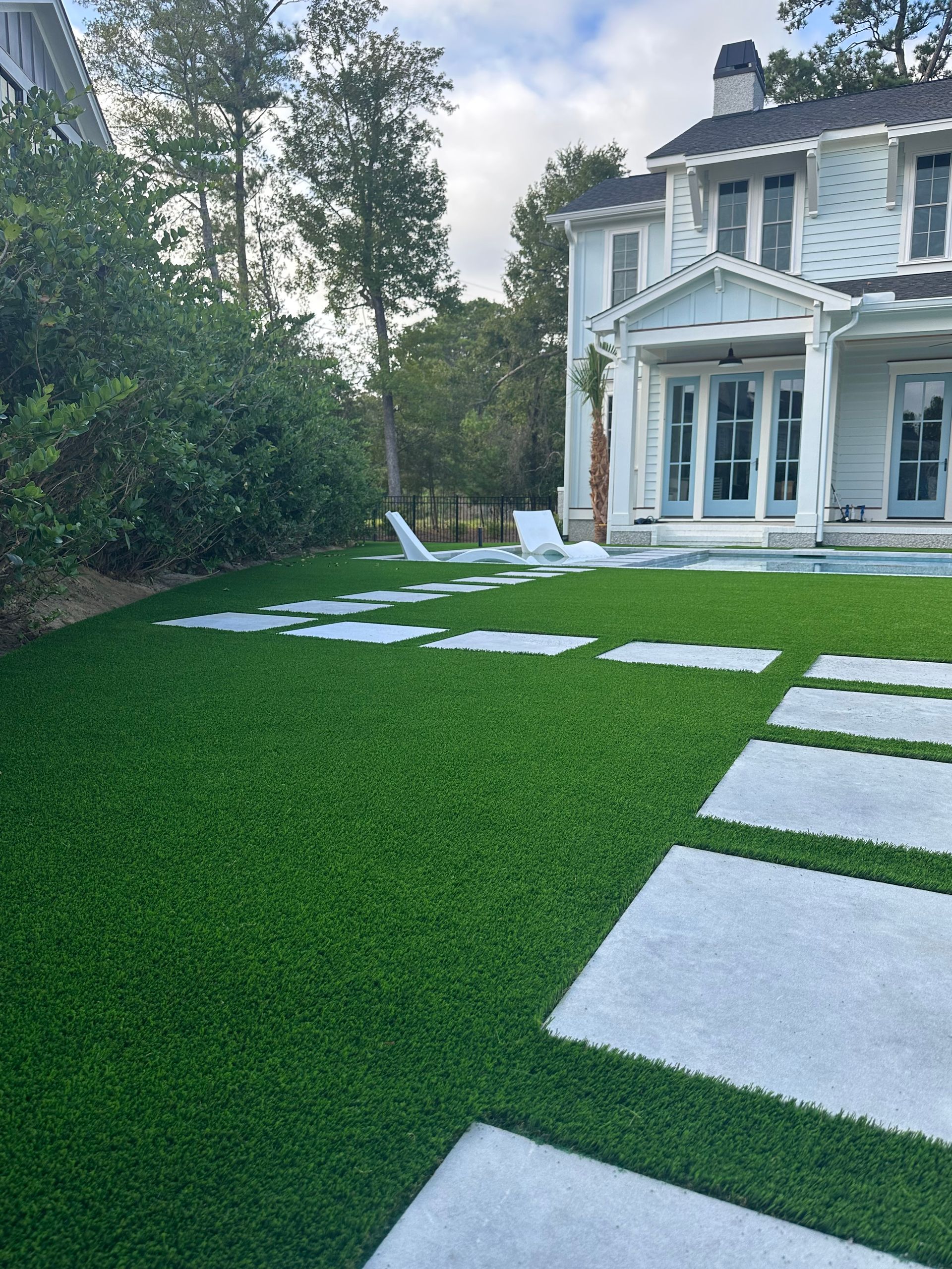 A backyard in Raleigh, NC, with artificial turf, stone pavers leading to a pool area, and a white two-story house in the background.