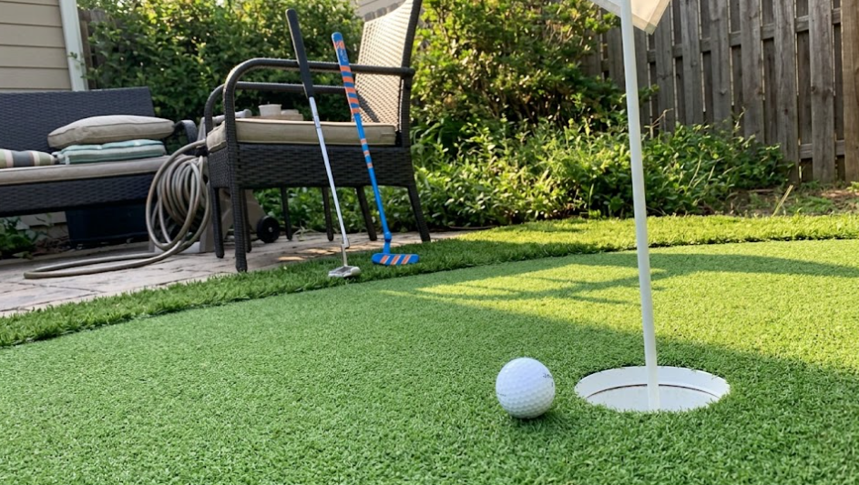 A white golf ball sits on a backyard artificial turf next to a hole with a flagstick; two putters stand nearby.