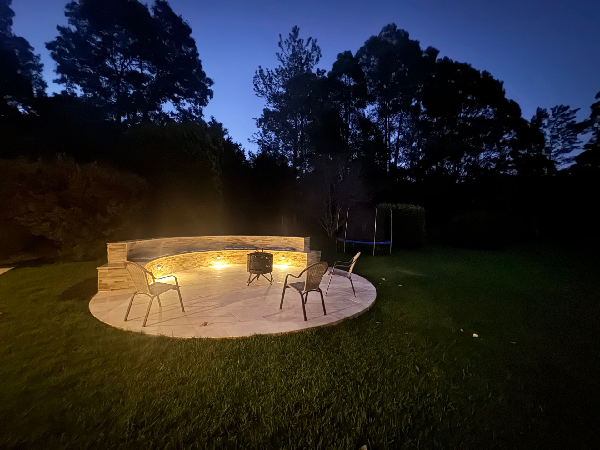 A stone patio with a curved fire pit wall and four chairs is illuminated at night in a grassy backyard.