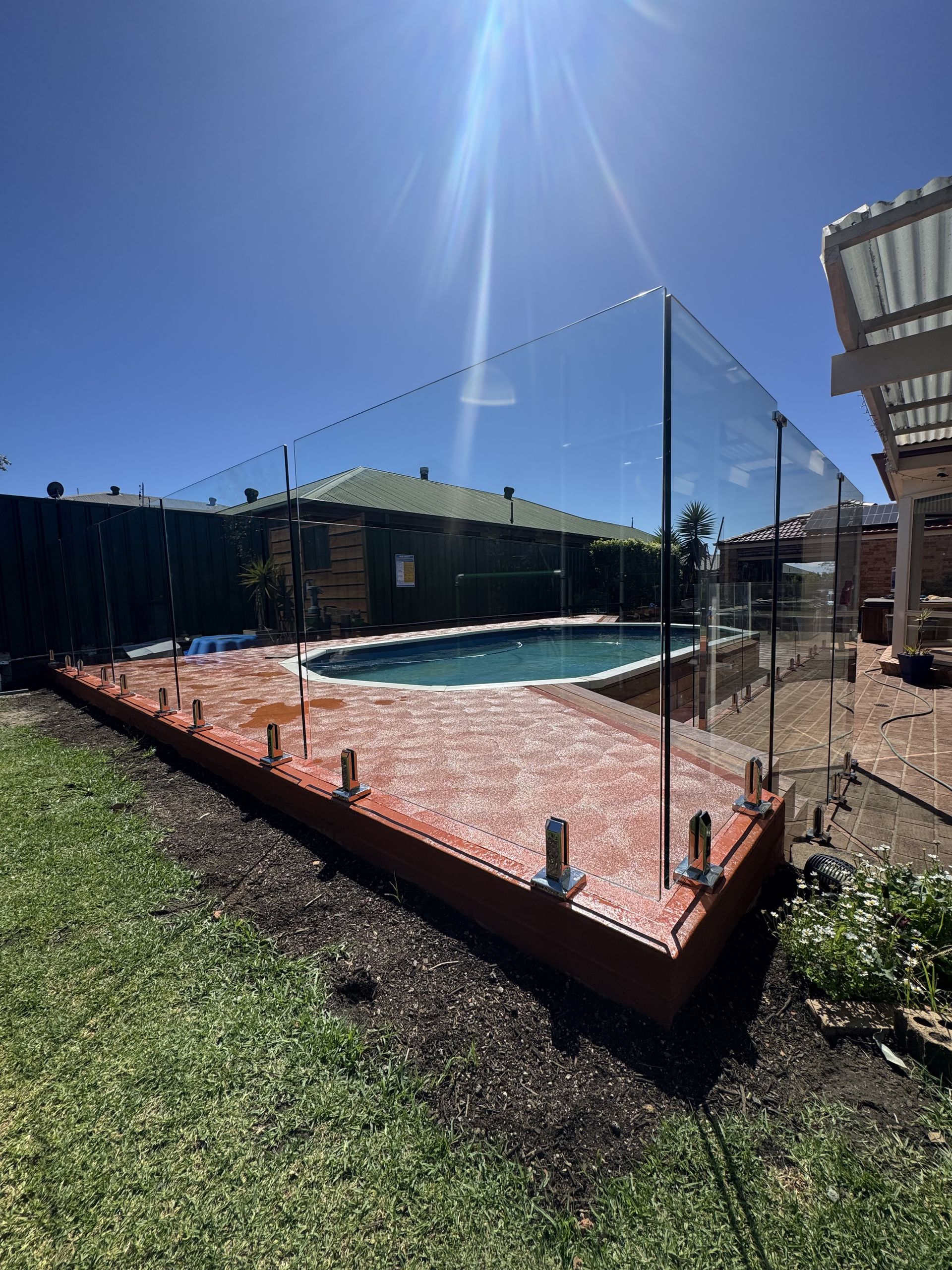 A backyard swimming pool area with a glass safety fence installed on a red brick deck under a bright, sunny sky.