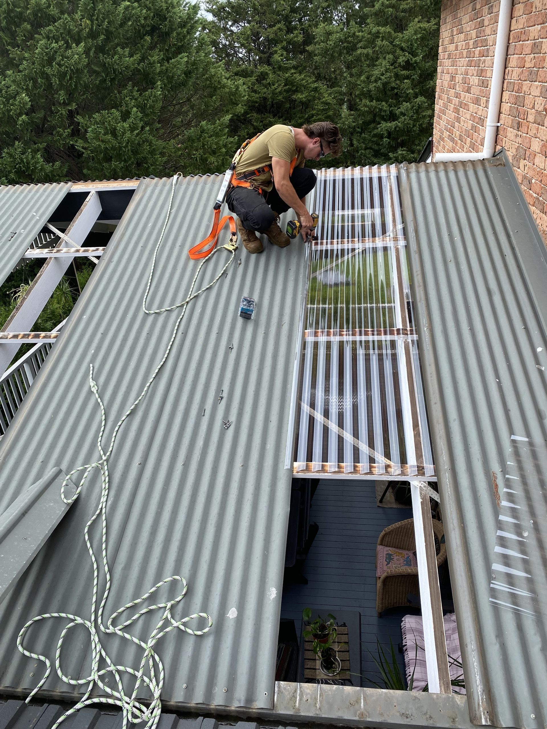 Person working on a corrugated metal roof, installing translucent panels. Safety harness and rope visible — CAW Services in Badagarang, NSW