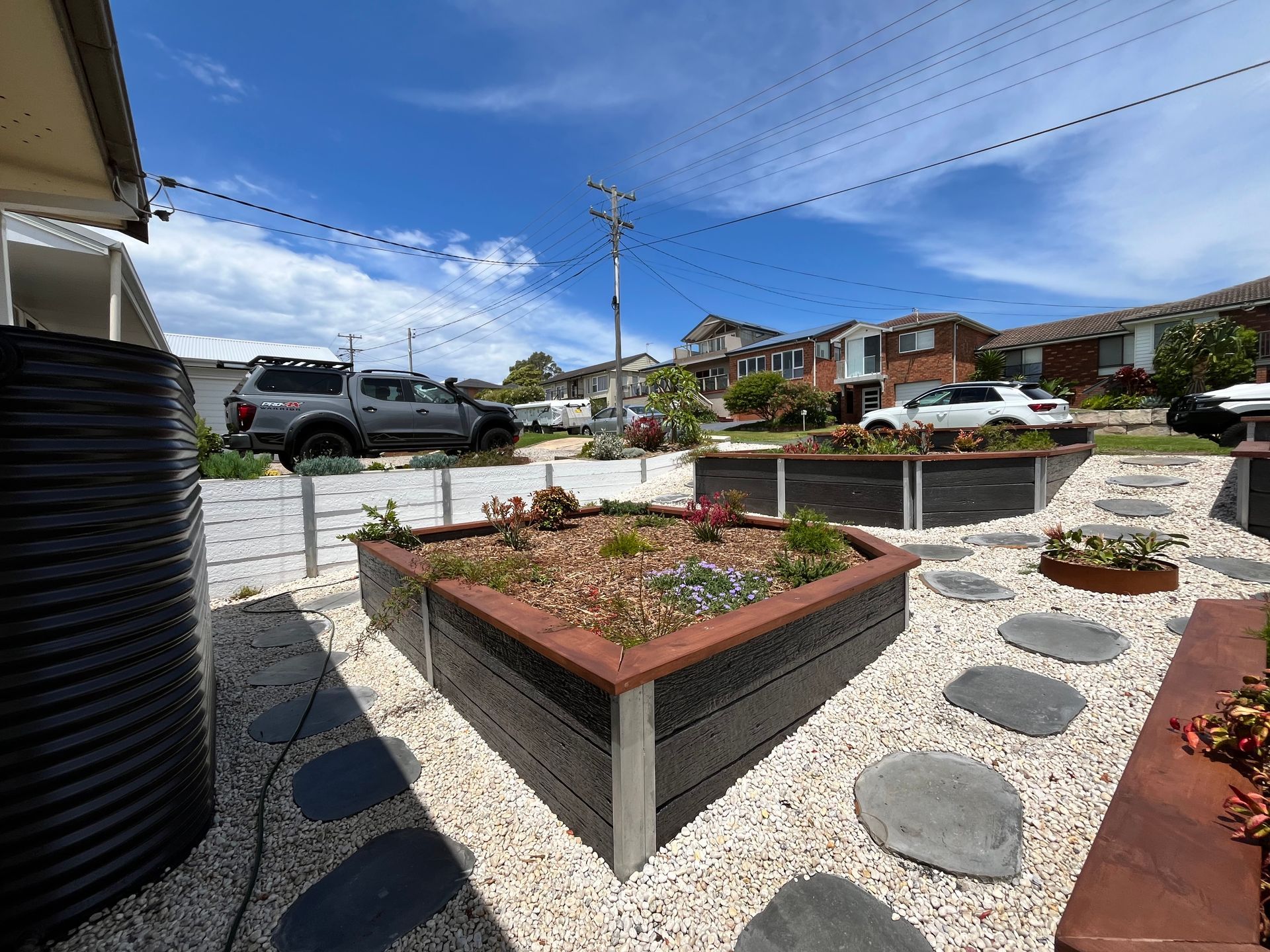 Front yard with raised garden beds, stepping stones, gravel, and a water tank. Cars and houses in background. — CAW Services in Badagarang, NSW