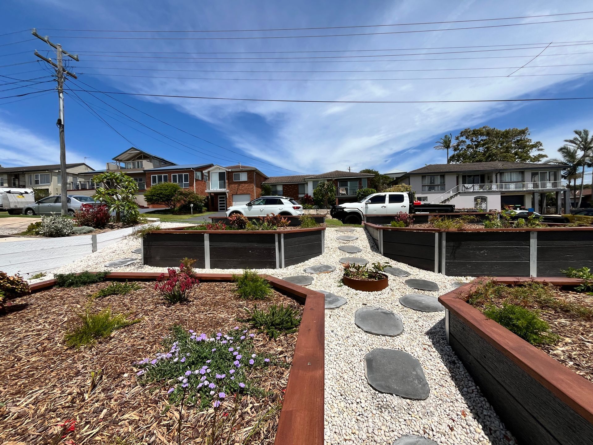 A landscaped garden path with stepping stones, light gravel, and raised wooden beds, with houses in the background.