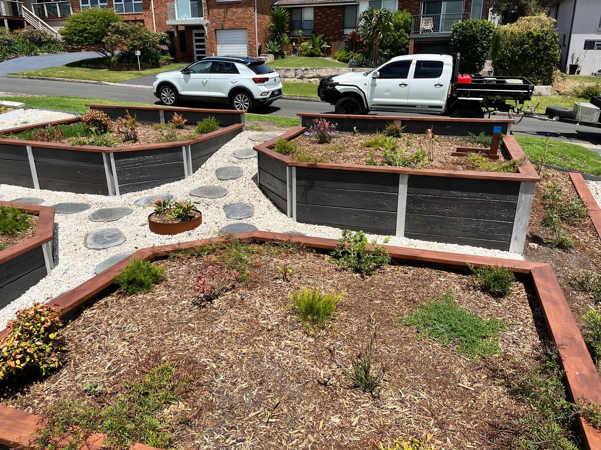 Raised garden beds with mulch and small plants are arranged on a gravel path near a suburban house and parked vehicles.