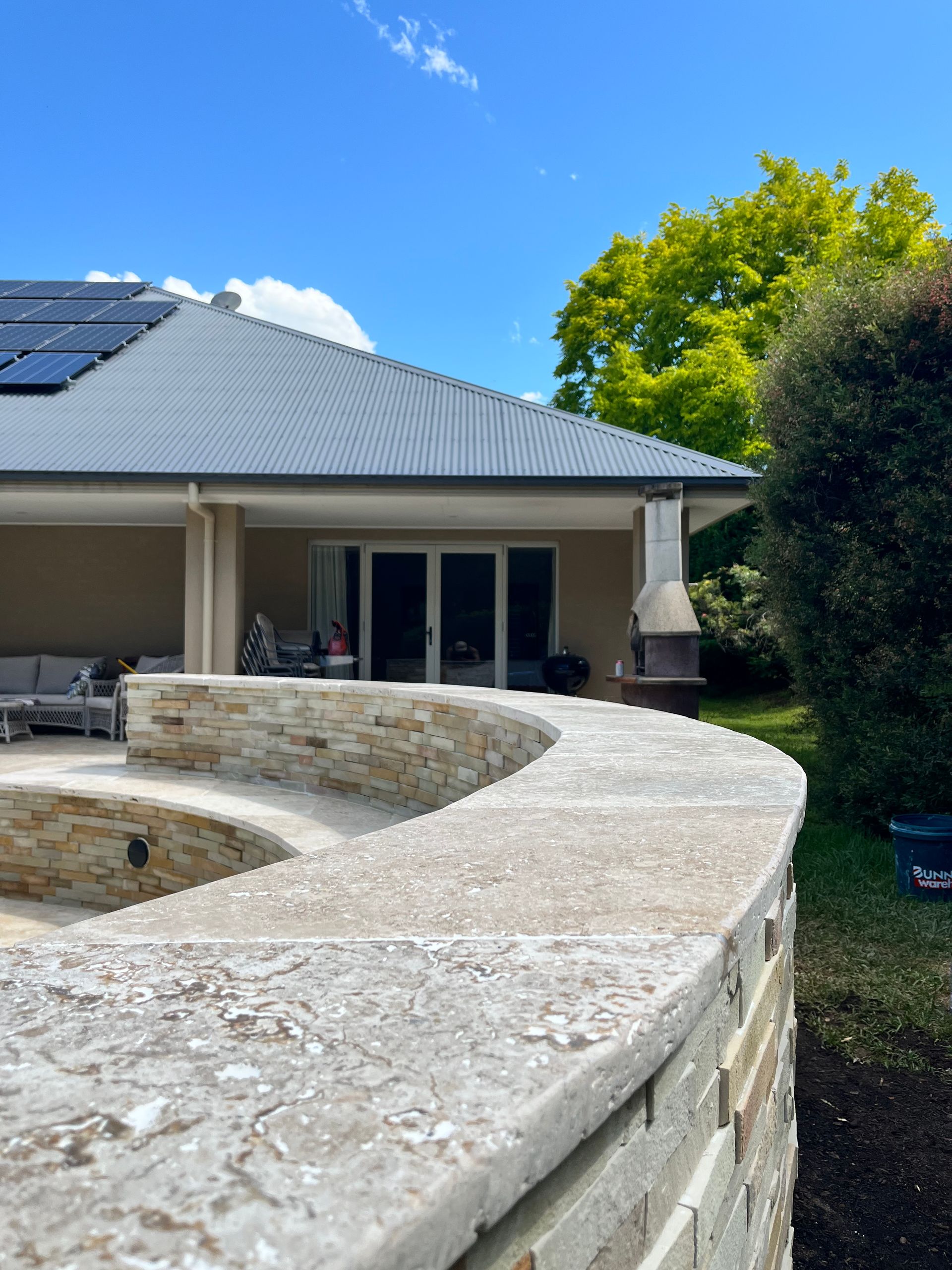 A curved stone retaining wall in a backyard patio area, set against a house with a gray roof under a blue sky.
