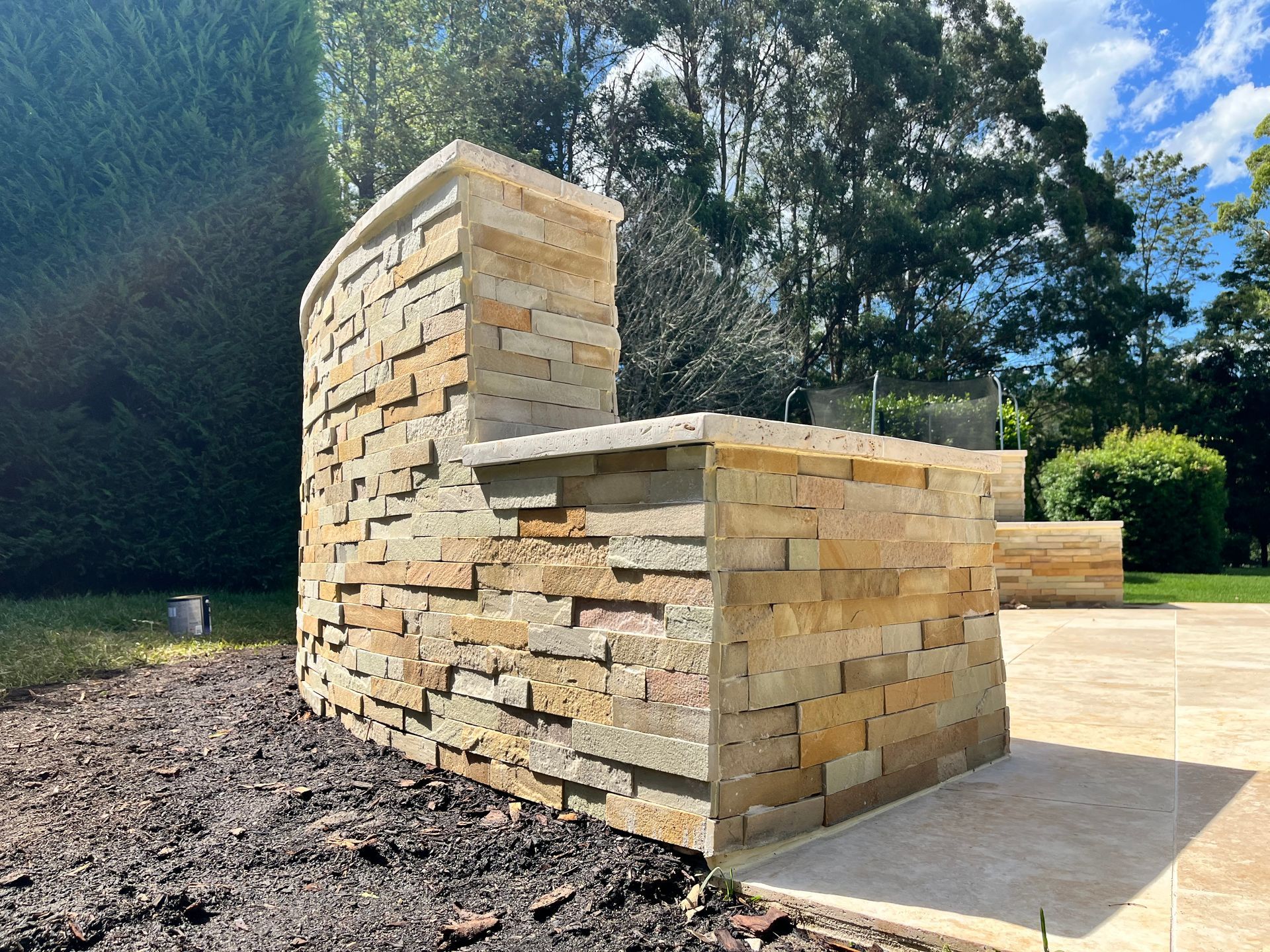 An outdoor stone counter with a raised backsplash, situated on a patio next to a garden bed with mulch.