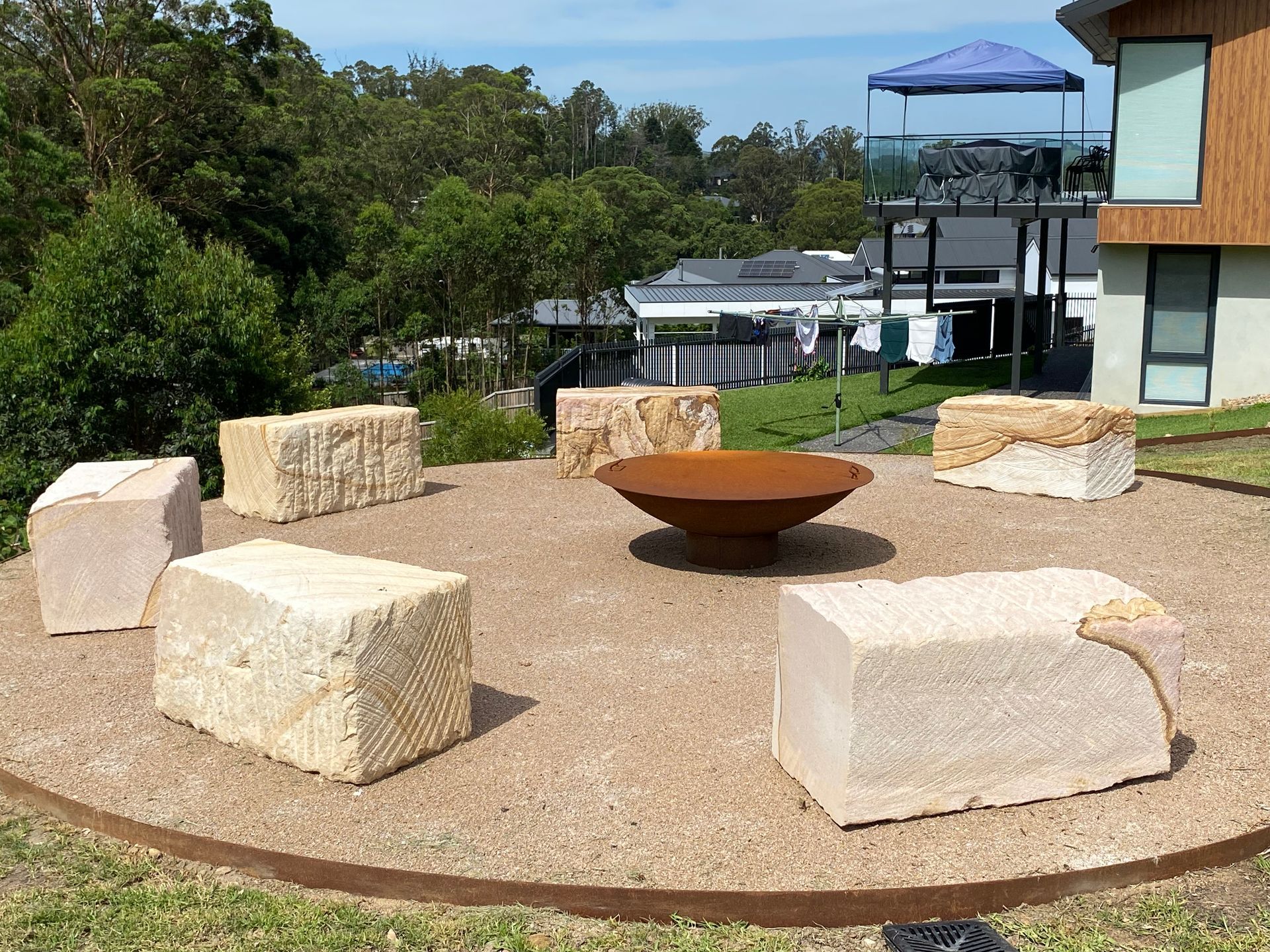 A circular gravel fire pit area with stone block seats arranged in a circle, set in a sunny, green outdoor space.