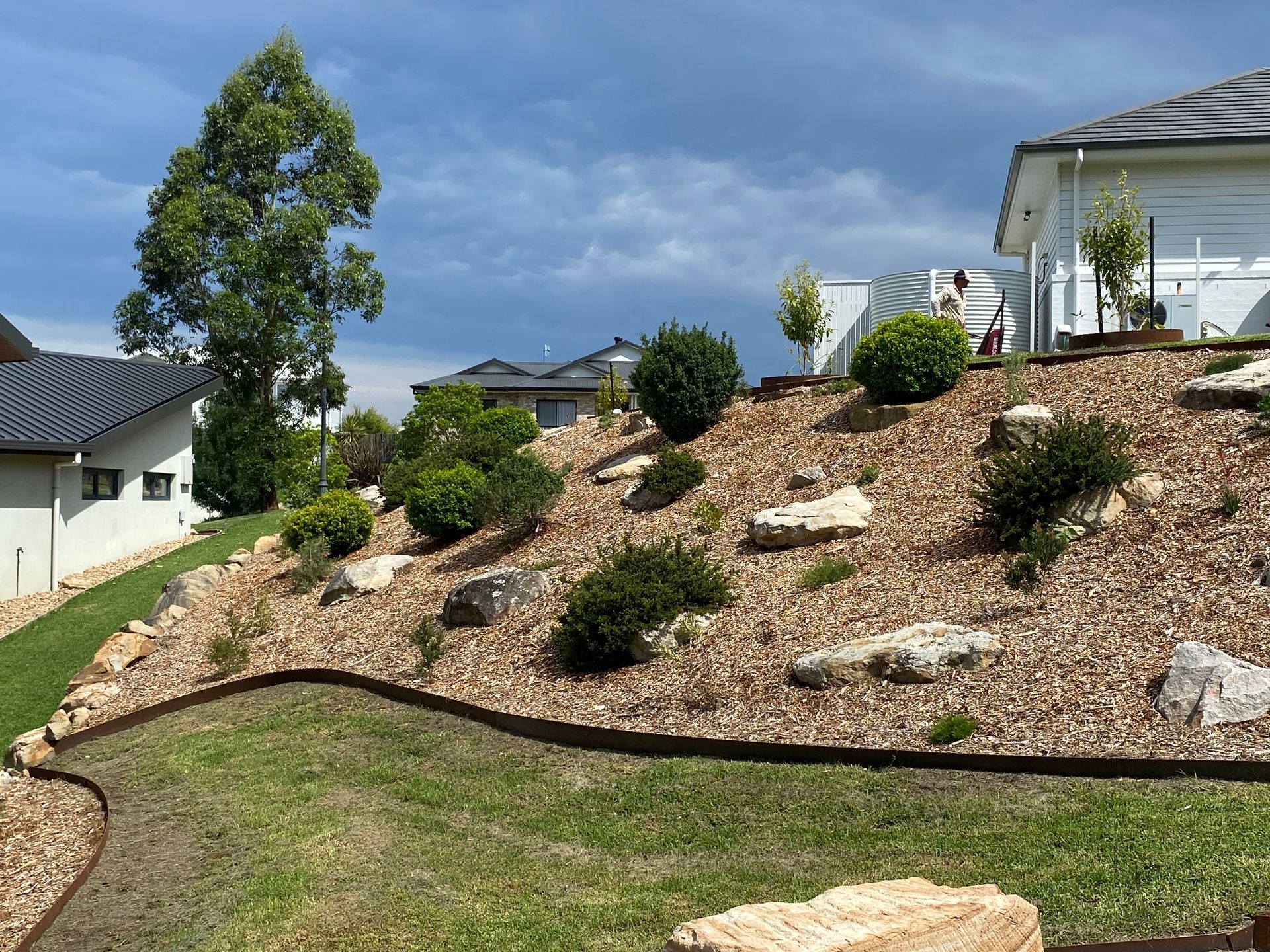 Landscaped hillside with mulch, shrubs, and rocks, under a cloudy sky, near houses — CAW Services in Badagarang, NSW