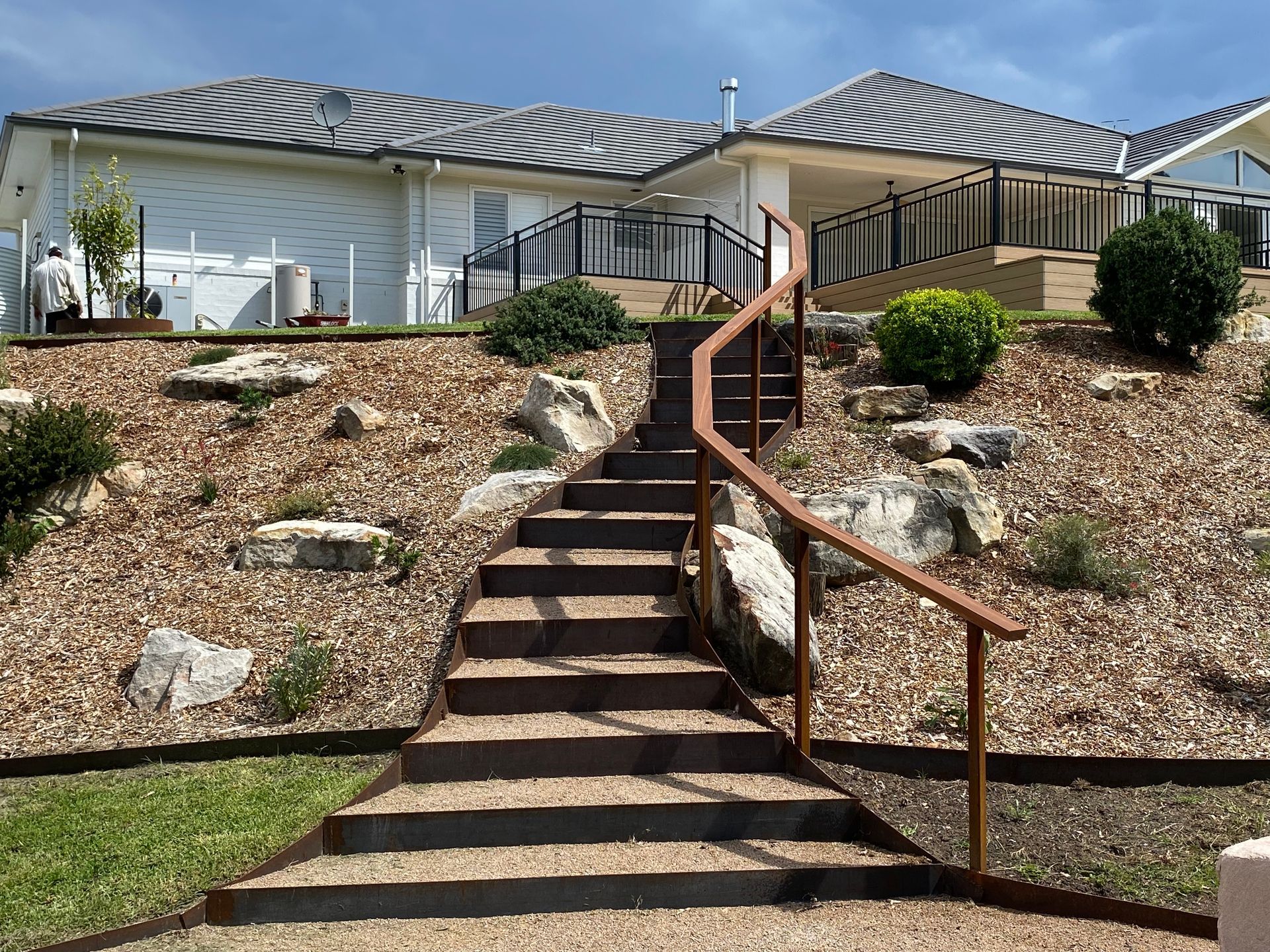 A front yard with raised garden beds and stone pathway. Cars parked on the street, houses in the background. — CAW Services in Badagarang, NSW