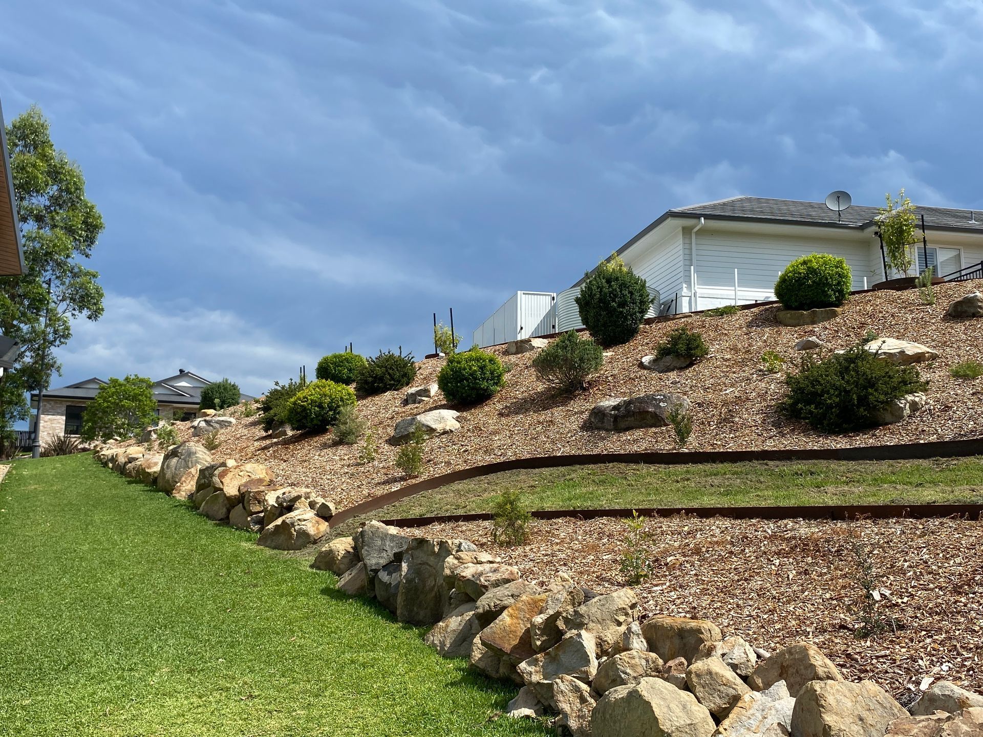 A landscaped hillside with a stone retaining wall, mulch, shrubs, and a house under a blue sky.