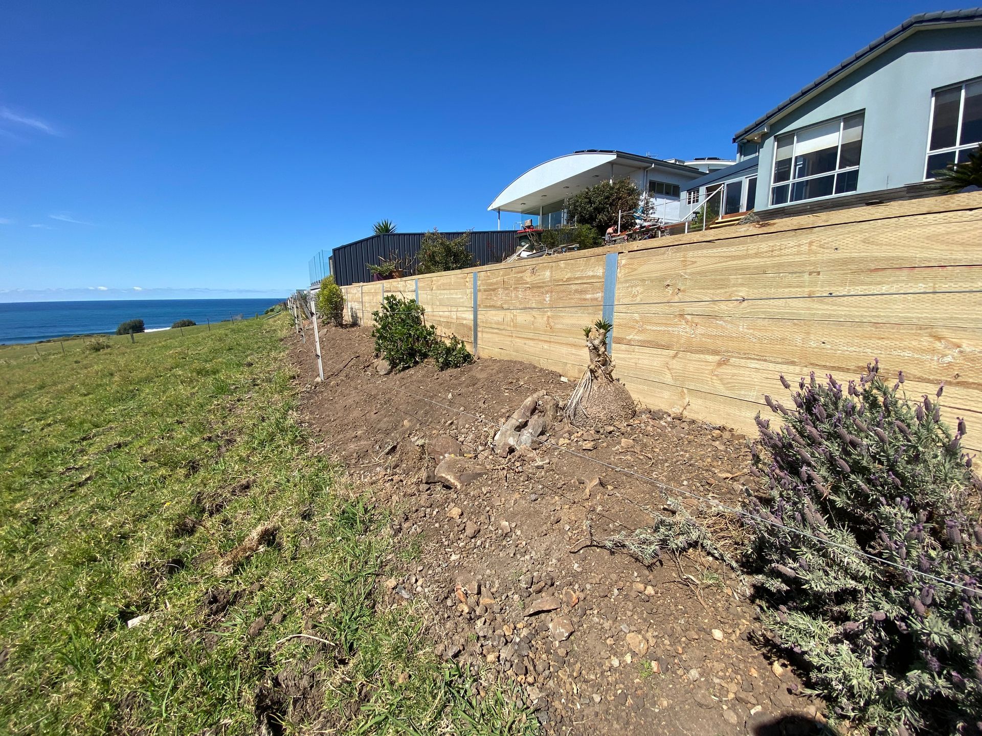 A wooden retaining wall borders a sloping grassy hill overlooking the ocean on a bright, sunny day.