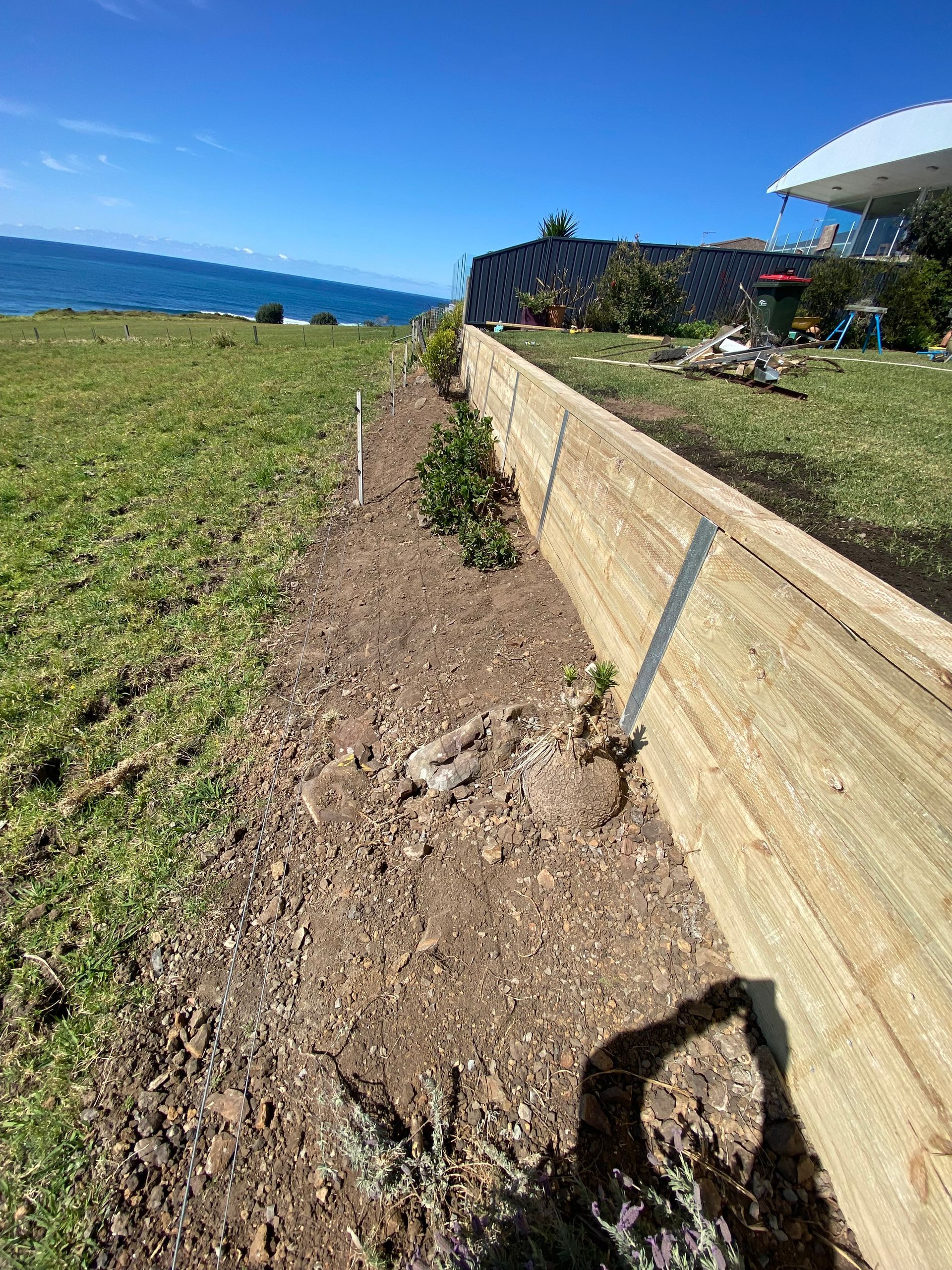 A timber retaining wall borders a mulch-filled garden bed, with a grassy lawn and the blue ocean in the background.