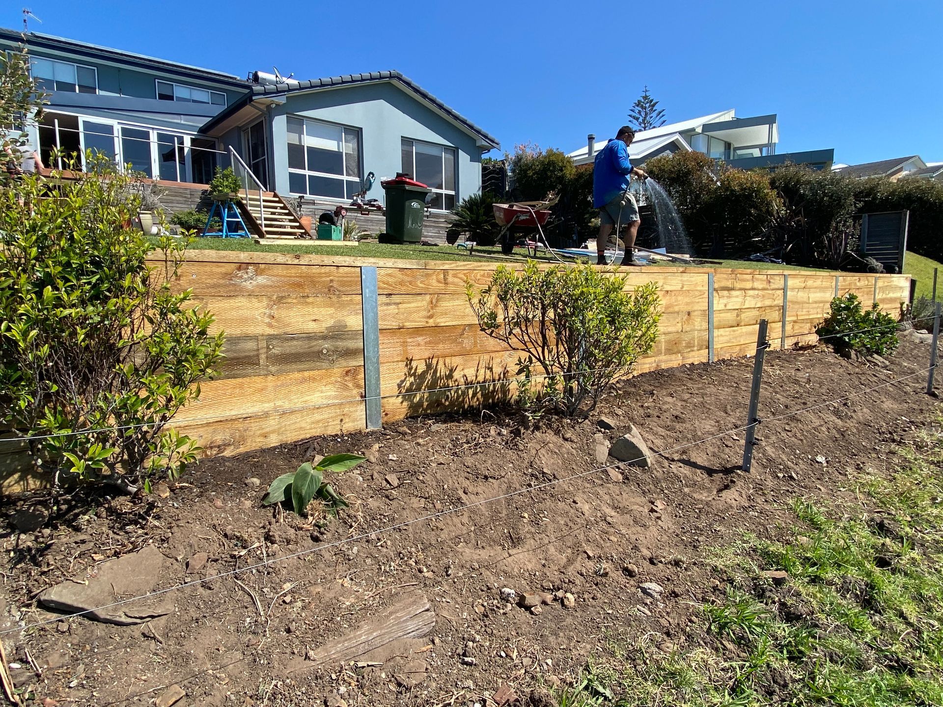 Man waters a new garden bed with a wooden retaining wall, set in front of a blue house. — CAW Services in Badagarang, NSW
