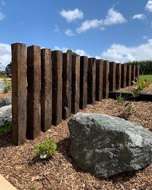 A row of dark, vertical timber posts stands in a garden bed with wood chip mulch and a large, gray landscape rock.