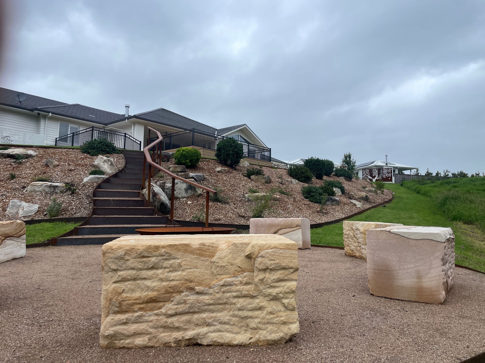 A stone-paved patio with large, rectangular sandstone seating blocks, featuring a staircase leading up to a modern house.