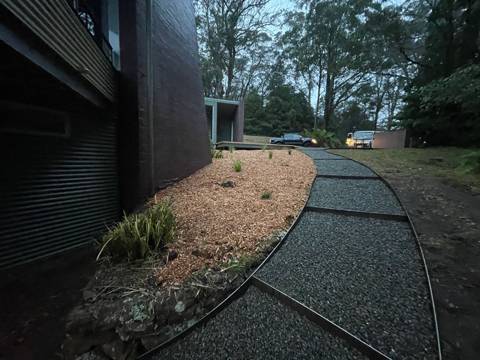 A path with grey gravel segments leads past a dark brick building and a mulched garden bed toward a wooded area.