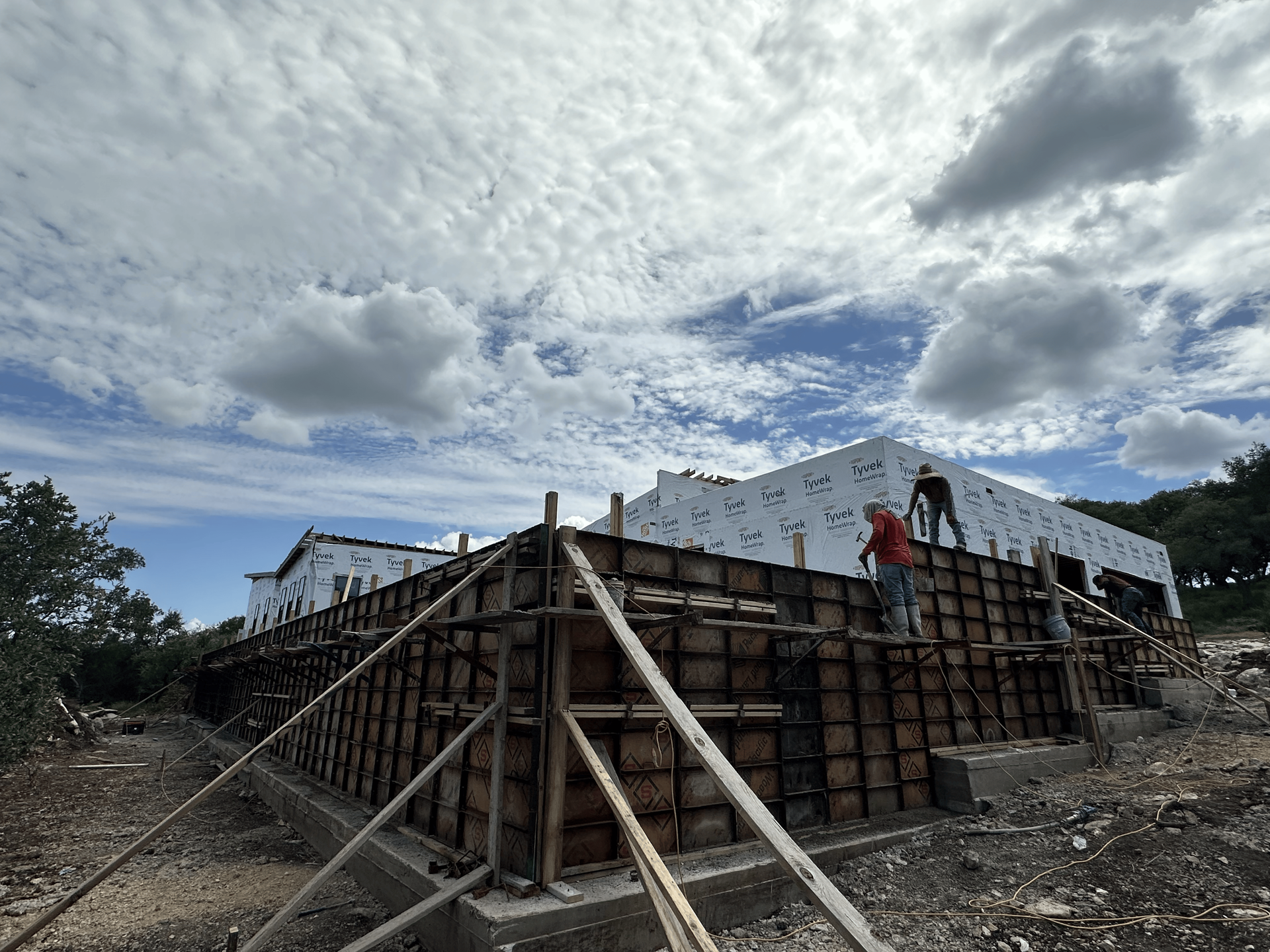 Construction site with workers, wooden framework for walls, cloudy sky.