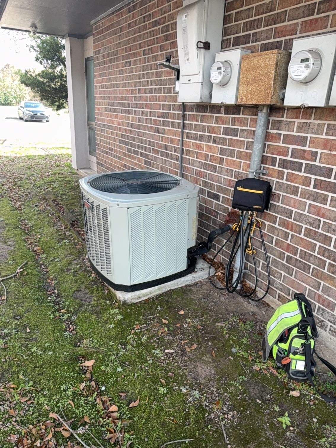 Outdoor air conditioner unit beside a brick house wall with utility meters and a backpack on the ground.