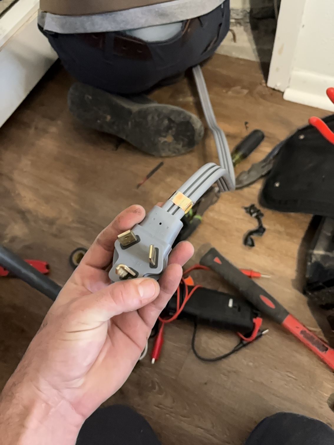 Hand holding a gray electrical connector with wires in a workshop, with tools on the floor nearby