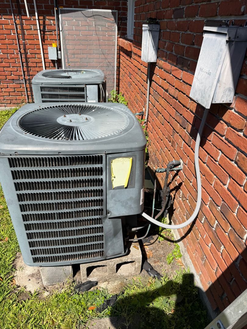 Two outdoor air conditioner units beside a brick wall with electrical conduit and sunlight shadows.