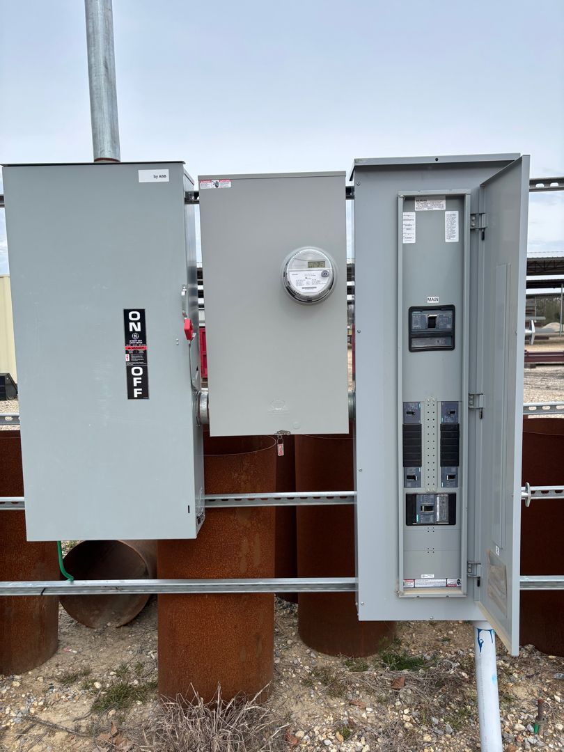 Three gray electrical meter boxes mounted outdoors on a wooden post beside a white pipe.