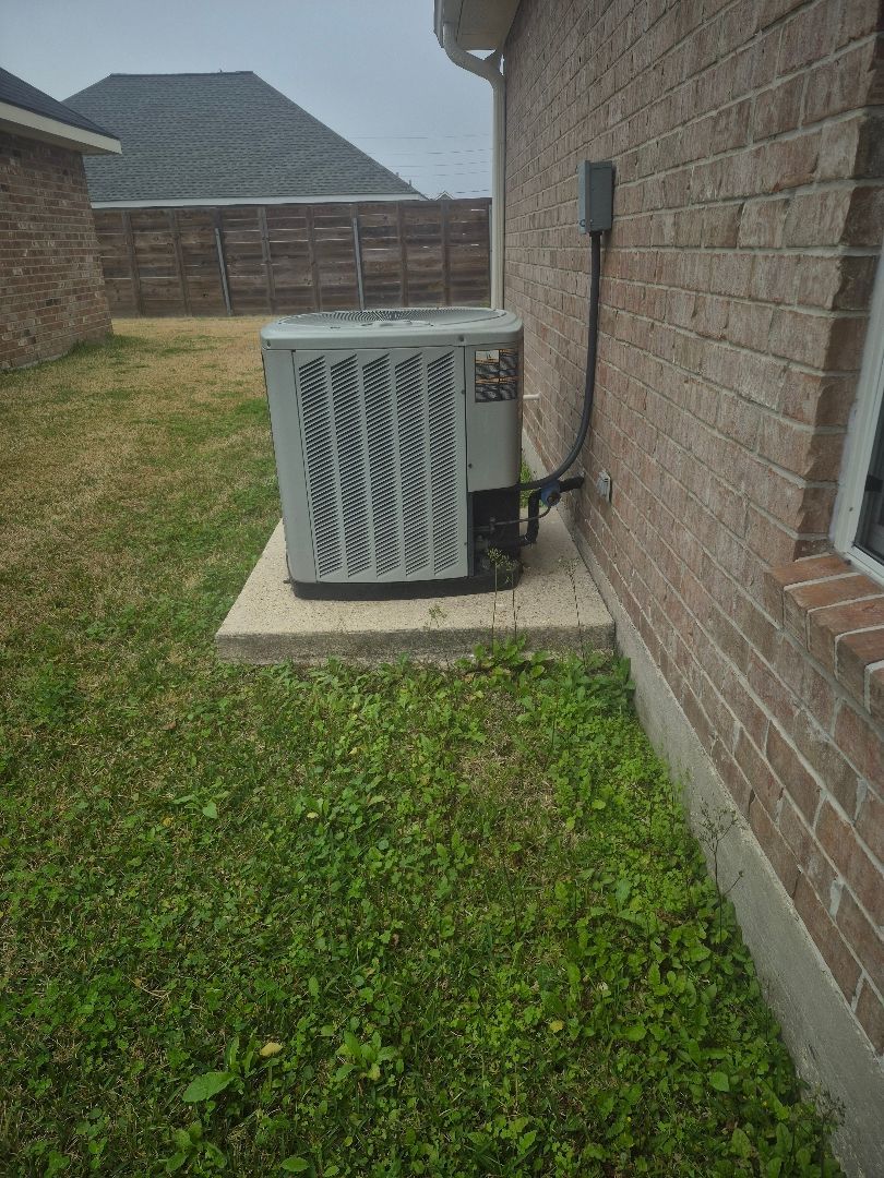 Outdoor air conditioner unit beside a brick house wall on a concrete pad, with grass and a fenced yard nearby