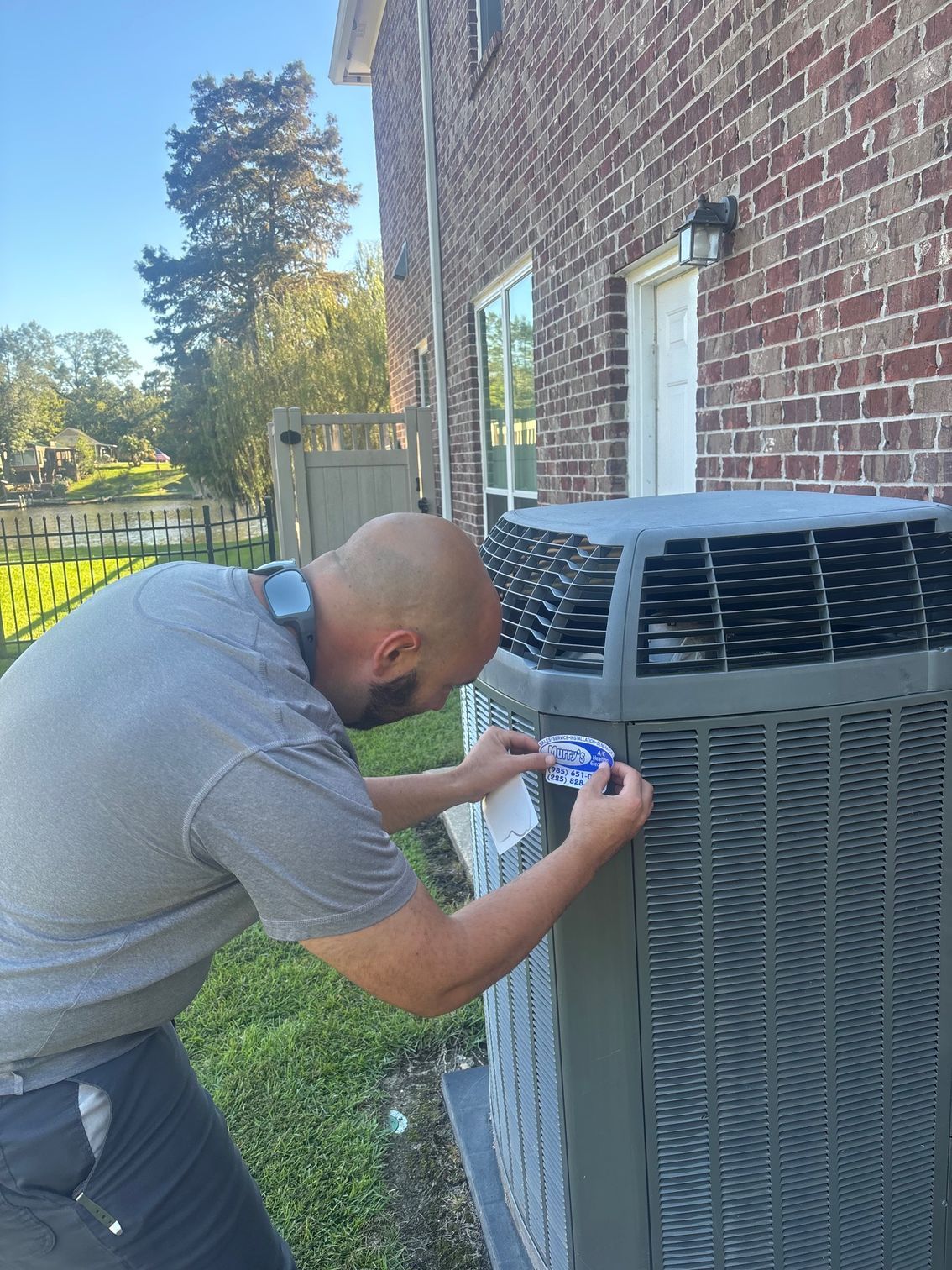 Man kneeling beside outdoor HVAC unit, tightening a bolt with a tool near a brick house and fenced yard