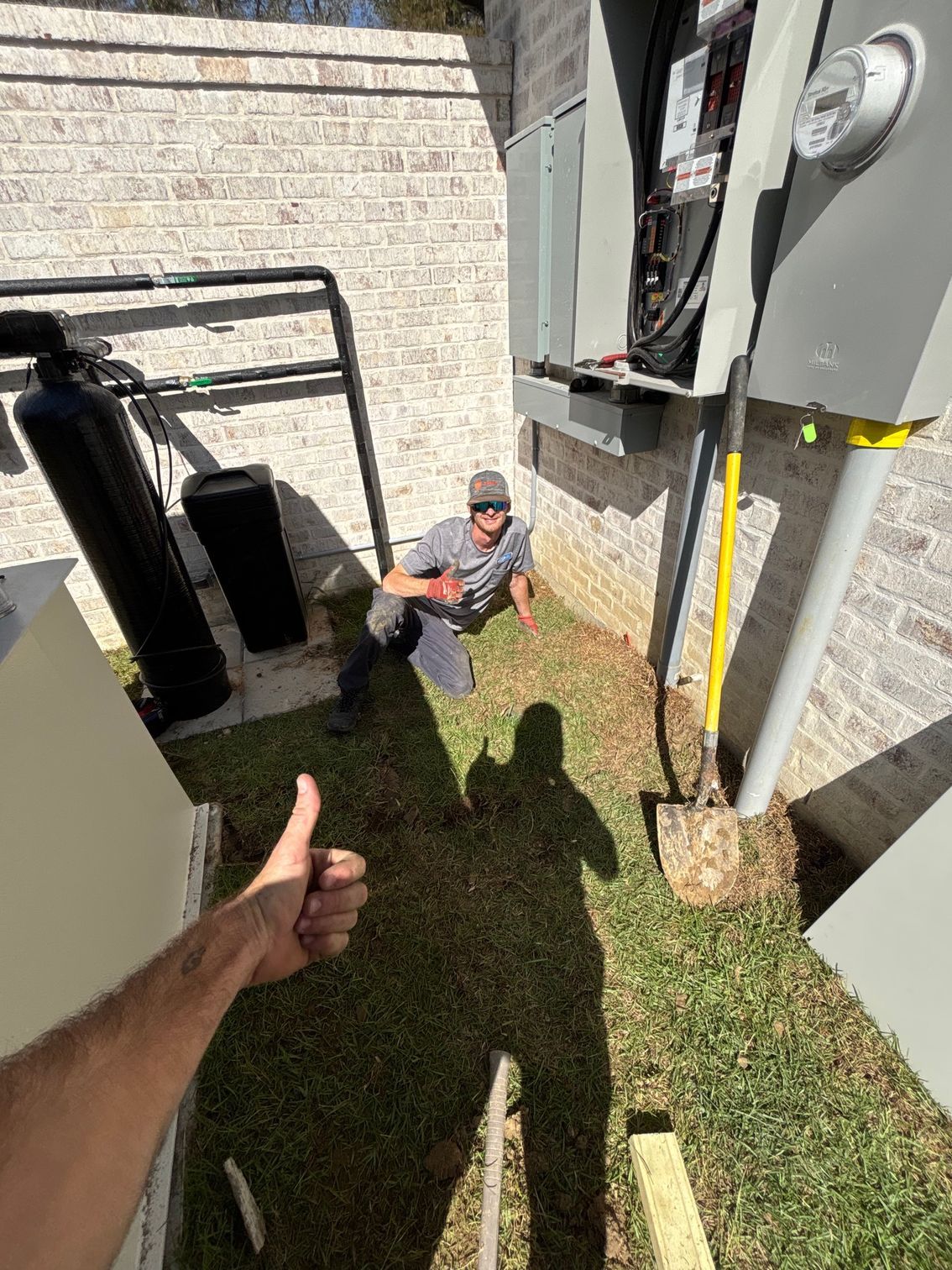 Person sitting on grass beside a house utility area, with a hand giving a thumbs-up in the foreground.