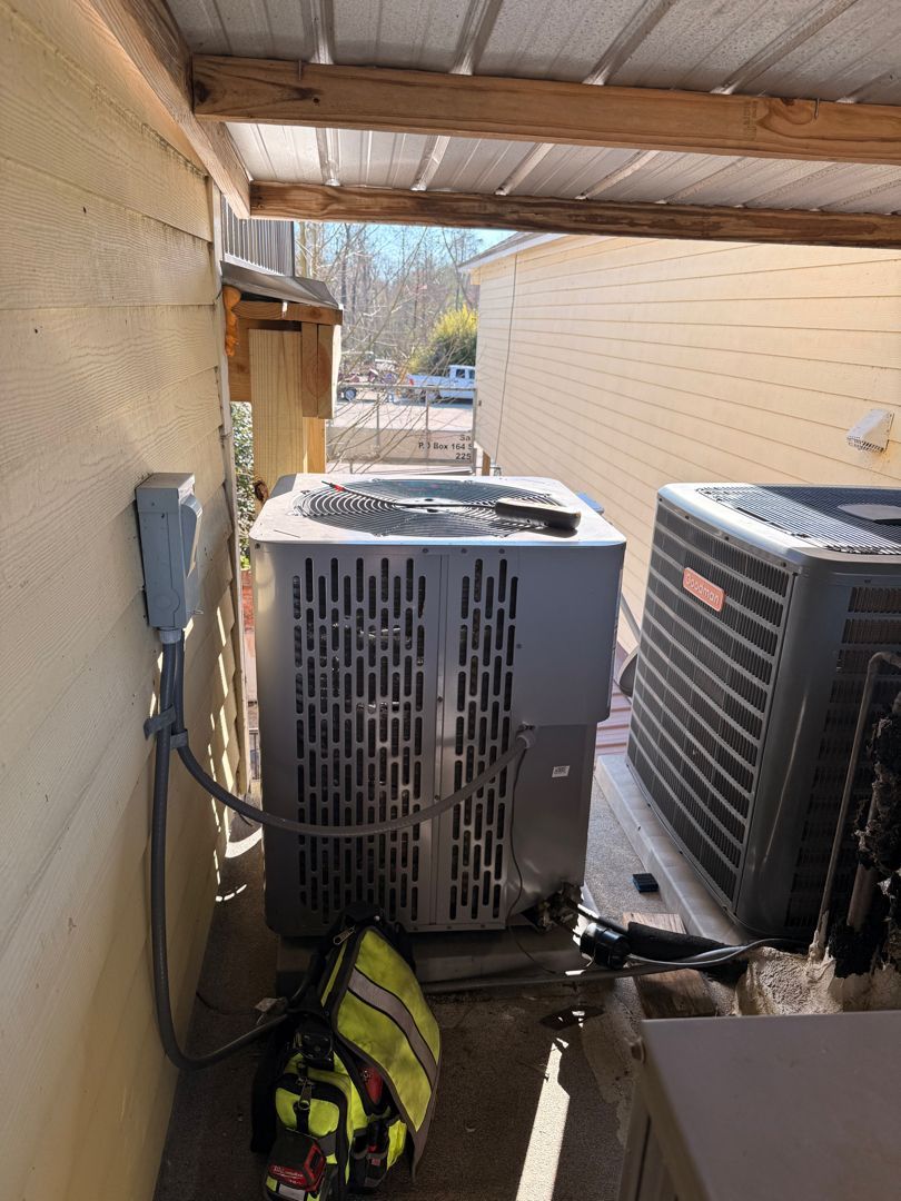 Outdoor HVAC units in a narrow covered side yard beside a stucco wall, with a firefighter bag on the ground.