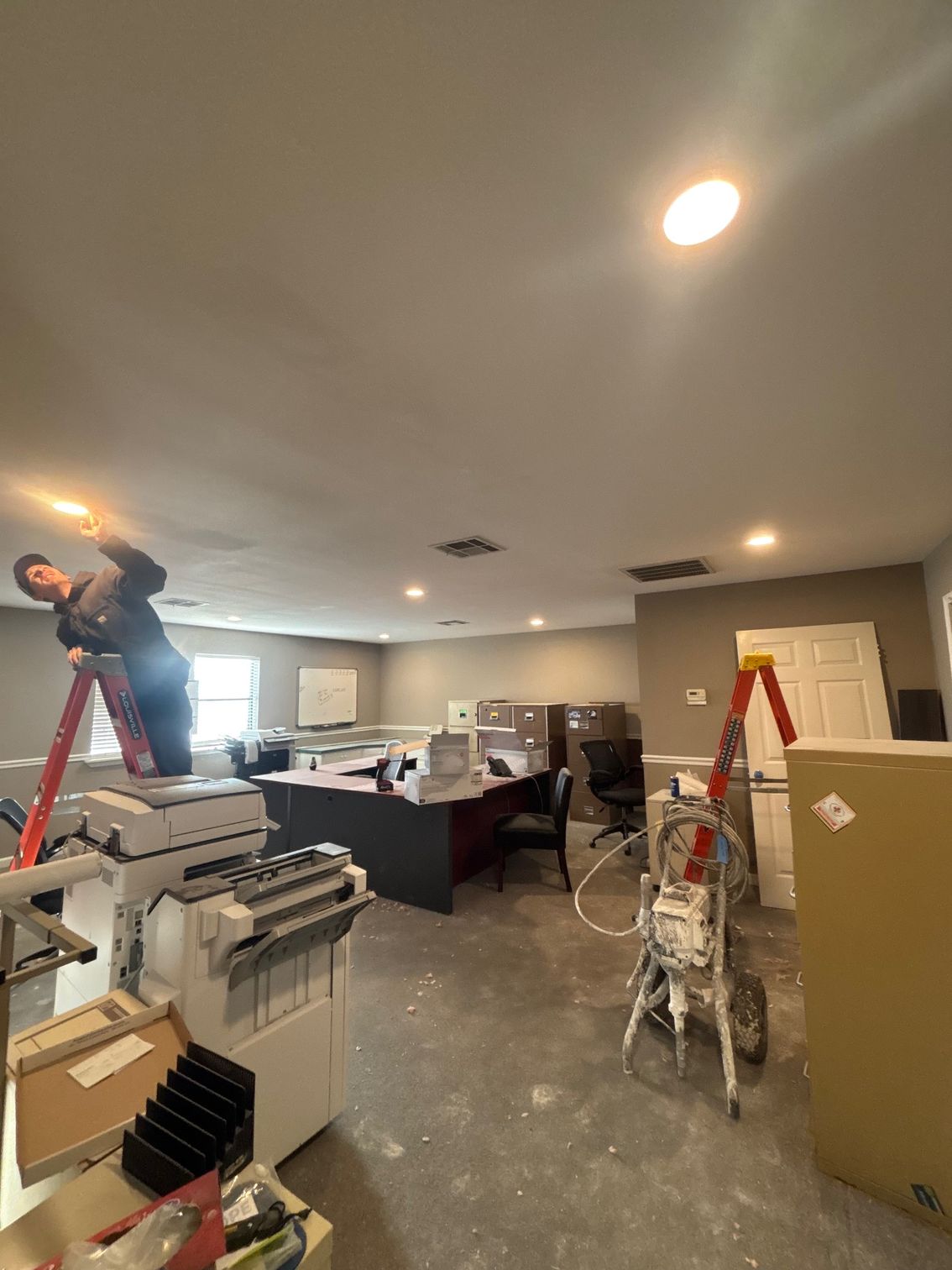 Workers renovating a room with ladders, tools, and drywall materials; one person works on the ceiling.