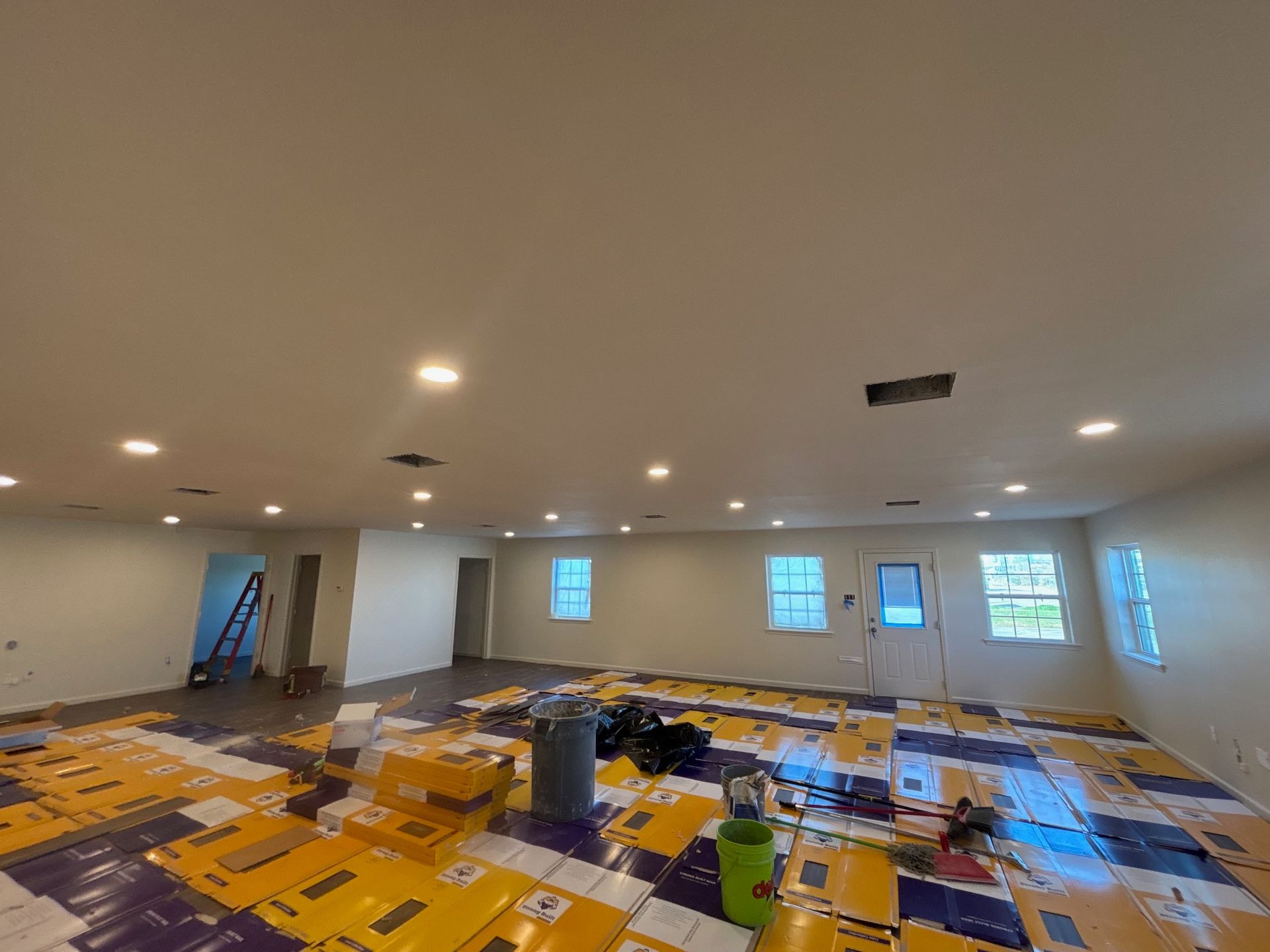 Large unfinished room with recessed lights, windows, and construction materials scattered across the floor.