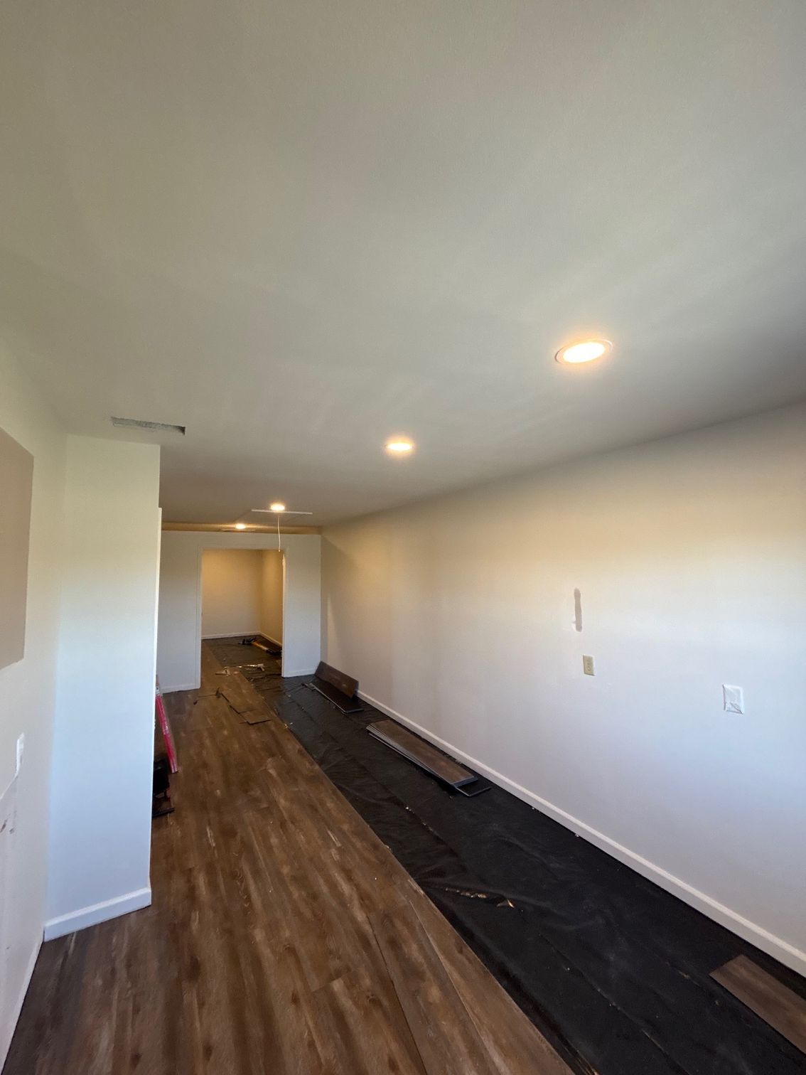 Long narrow hallway with wood flooring, white walls, recessed lights, and a black floor strip.