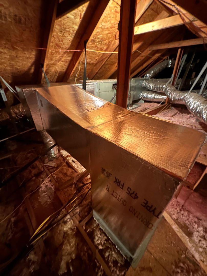 Attic HVAC ductwork and insulation beneath wooden rafters, lit by warm light.
