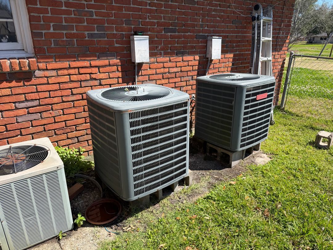 Three outdoor HVAC units beside a brick house, with grass and a small bucket nearby.