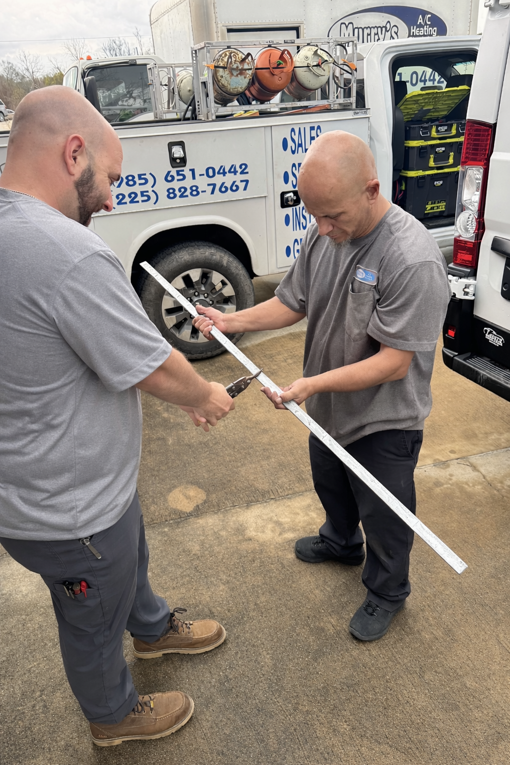 Two workers in gray shirts inspect a long white pole beside a service van.